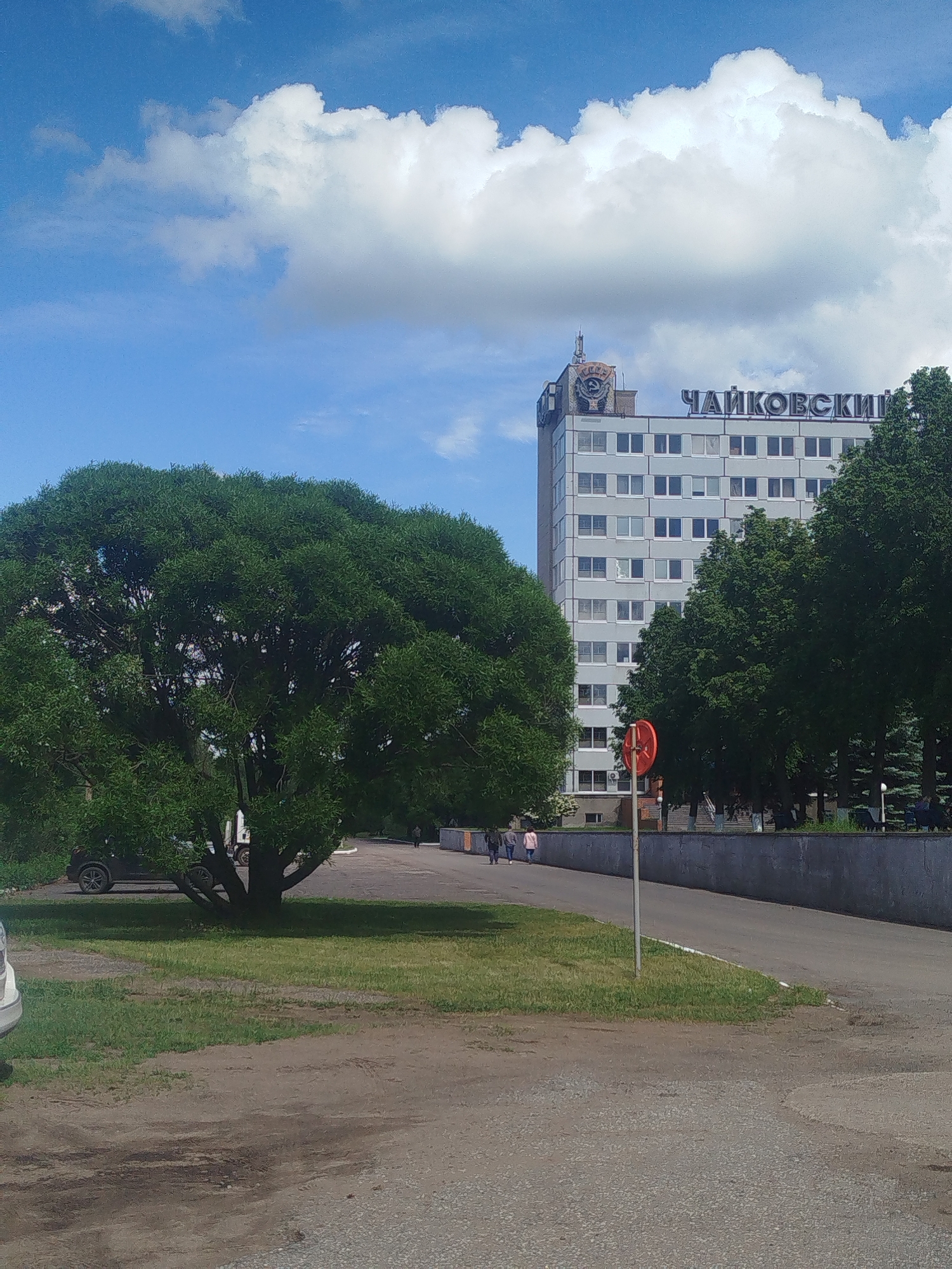 The image depicts a street view in what appears to be the city of Tchaikovsky, Russia, as indicated by the Cyrillic text "ЧАЙКОВСКИЙ" on the top of a multi-story building. The building, with a grey facade and numerous windows, features a Soviet-era emblem on its roof.
In the foreground, there is a large, bushy tree to the left, providing shade to a grassy area. A road divides the foreground from the background, with a red and white traffic sign visible on the right side of the road. Several individuals are walking along the sidewalk in the mid-ground. A low concrete wall runs behind the sidewalk.
The sky is blue with scattered white clouds. Several cars are parked in the vicinity of the tree on the left. The overall setting seems to be a somewhat urban environment with a mix of greenery and infrastructure.