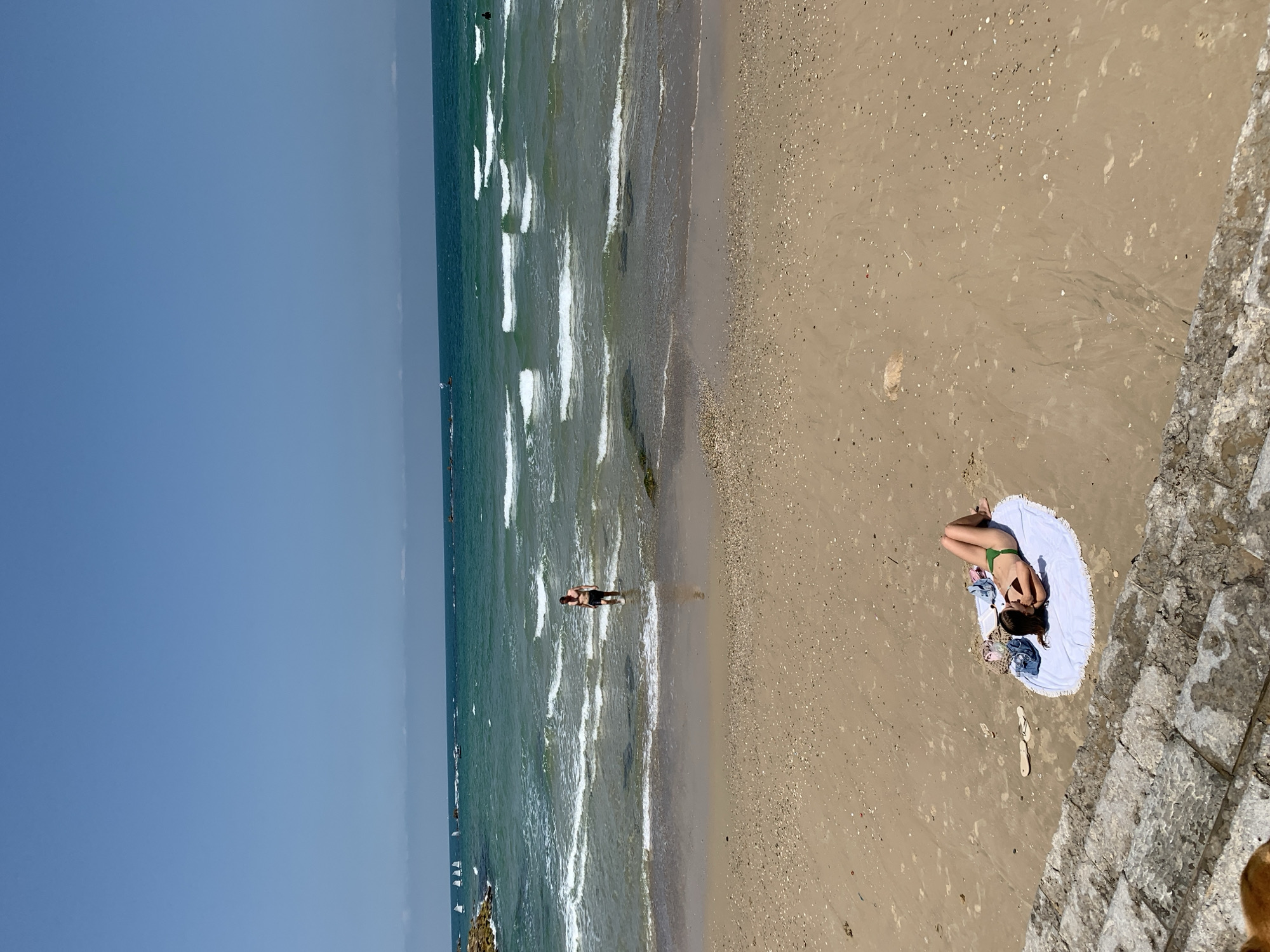 Here's a forensic description of the image:
**Location:** The image appears to be taken at a beach. The city is Tel-Aviv, Israel, based on typical scenery of Tel-Aviv beaches.
**Environment:** A bright, clear day at the beach. The scene includes the Mediterranean Sea, sandy beach, and a stone wall or structure at the edge of the beach.
**People:**
* A woman lying on a round white towel on the sand wearing a green bikini.
* A person stands in the water, possibly swimming or wading.
* Other individuals can be seen in the distance within the sea.
**Objects:**
* White round towel.
* Swimsuit.
* Sandals.
* Sea
* Stone construction
**Actions/Interactions:**
* Woman sunbathing on the beach.
* Other person stands in the water.
**Overall Impression:** The scene depicts a sunny day at the beach. There is a woman relaxing on the beach, and someone else is enjoying the water. There are sail boats in the water. The image suggests a typical beach scene in Tel-Aviv.