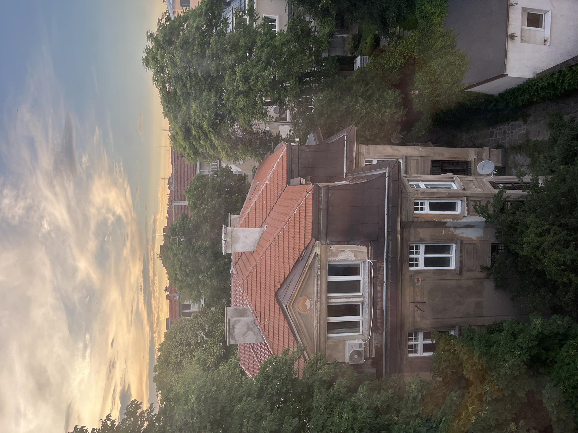 This is an aerial view of a weathered, old building with a red tiled roof, located in the city of Sofia, Bulgaria. The building appears to be a residential house, possibly from the early 20th century, with architectural details such as ornate window frames and a triangular pediment above a large window. The facade of the building is a faded ochre color, with visible signs of age and deterioration.
The house is surrounded by lush green trees, obscuring parts of the structure and creating a sense of depth and density in the urban landscape. Beyond the immediate building, other residential buildings with red roofs and balconies are visible, interspersed with more trees, suggesting a neighborhood setting. The sky above is filled with soft, diffused light, with clouds tinged with the warm hues of either sunrise or sunset. There are no people visible in the image, and the scene is quiet and still. The overall impression is one of a peaceful, albeit slightly aged, urban setting at dusk or dawn.