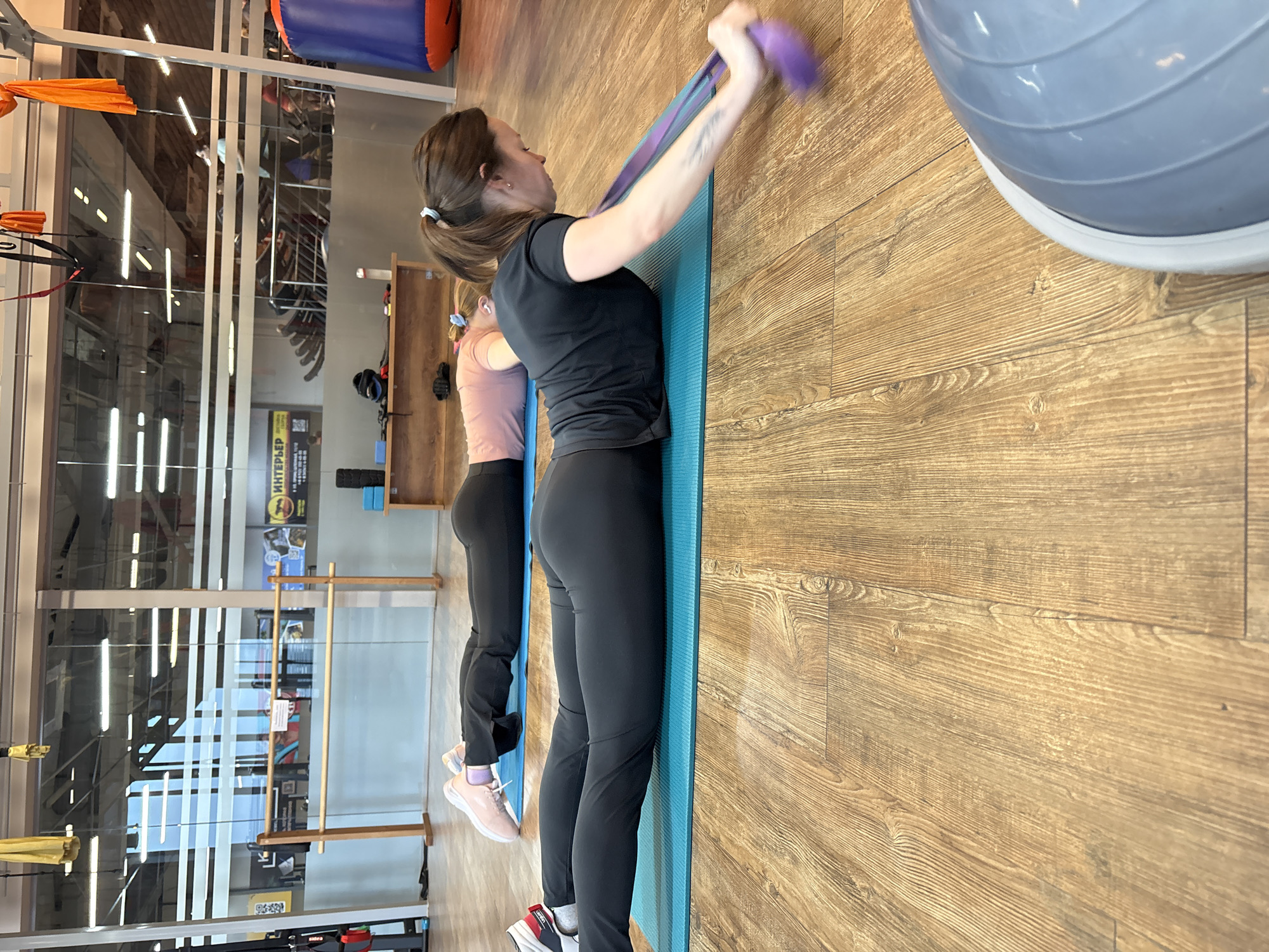 Two women are exercising in a fitness facility, likely in the city of Chaikovsky, Russia. The main subject is a woman in the foreground dressed in black athletic attire, including a black t-shirt and black leggings, with her hair pulled back in a ponytail. She is standing on a blue yoga mat and holding a purple resistance band stretched above her head. The second woman, further in the background, is also on a blue yoga mat and dressed in athletic clothes. She is closer to a glass wall showcasing exercise equipment, suggesting this is a gym setting. A large grey exercise ball is partially visible in the upper right corner of the frame. The floor is wood laminate. A sign in Russian is visible through the glass wall, confirming the location is Russia.