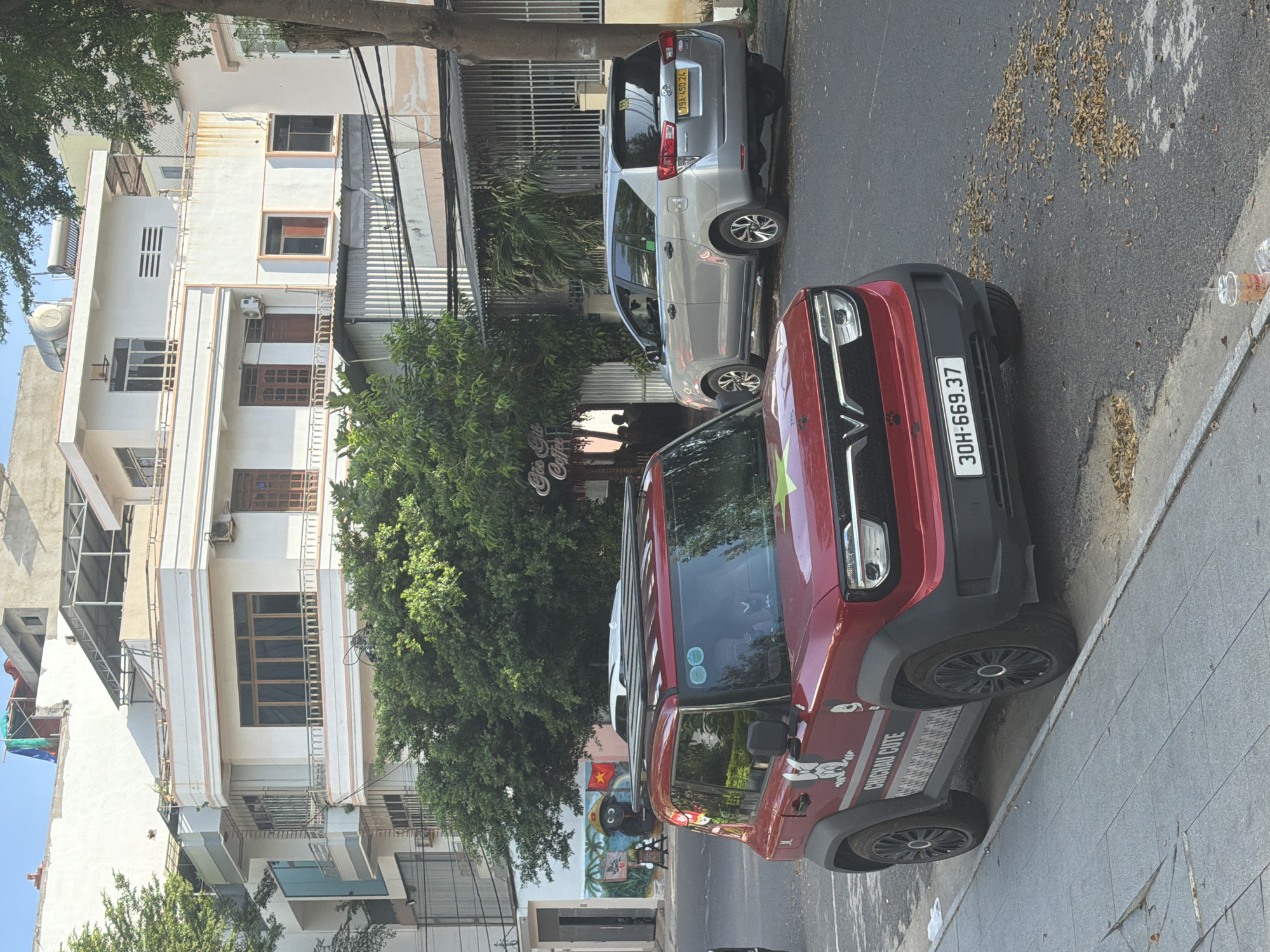 The image depicts a street scene in Bac Nha Trang Ward, Vietnam. In the foreground, a red electric vehicle is parked at the side of the road, featuring a rack on its roof and decals on its doors, including the text "CHICHAI CUTE" and a cartoon dog graphic. To the right of the red vehicle, a silver minivan is also parked on the street. Behind the parked vehicles, a building with multiple stories is visible, adorned with white and cream-colored facades, balconies with railings, and dark-framed windows. Trees with dense green foliage line the street, obscuring parts of the buildings. A sign for "Goc Gia Coffee" is also visible. The pavement in the foreground shows some discoloration and scattered dry leaves. The sky is clear and blue.