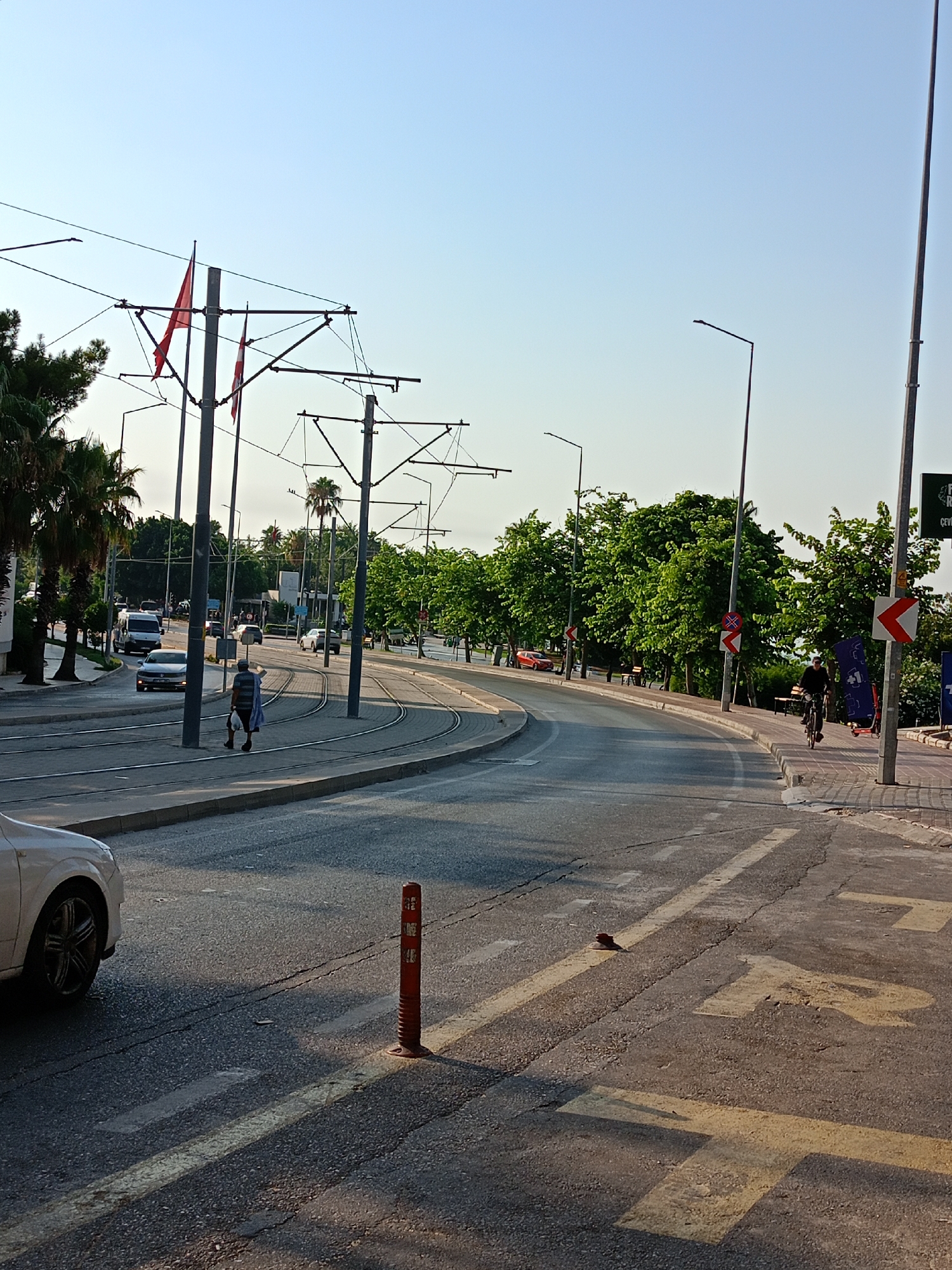 The image captures a street scene in Antalya, Turkey, likely near a tram line, given the presence of overhead wires and tracks. The photograph is taken from a slightly elevated perspective, looking down a curved road lined with palm trees and lush green foliage on the right. The sky is clear and a light blue.
In the foreground, a white car is partially visible on the left. The road surface shows yellow markings and a red traffic cone. Further down the road, a person is walking with shopping bags, dressed in a dark shirt and shorts, and a light blue garment draped over their shoulder. Tram tracks are visible on the road, curving to the right.
Several vehicles are present on the road, including a white van and other cars. There are also streetlights and poles supporting the tram overhead wires. On the right side of the road, a person is cycling and another person is standing near a bicycle. Traffic signs, including directional arrows and a circular red sign with a diagonal bar, are visible.
The overall atmosphere suggests a sunny day in a city environment, with elements of urban infrastructure and everyday life. The presence of palm trees indicates a warm climate, consistent with Antalya's location.