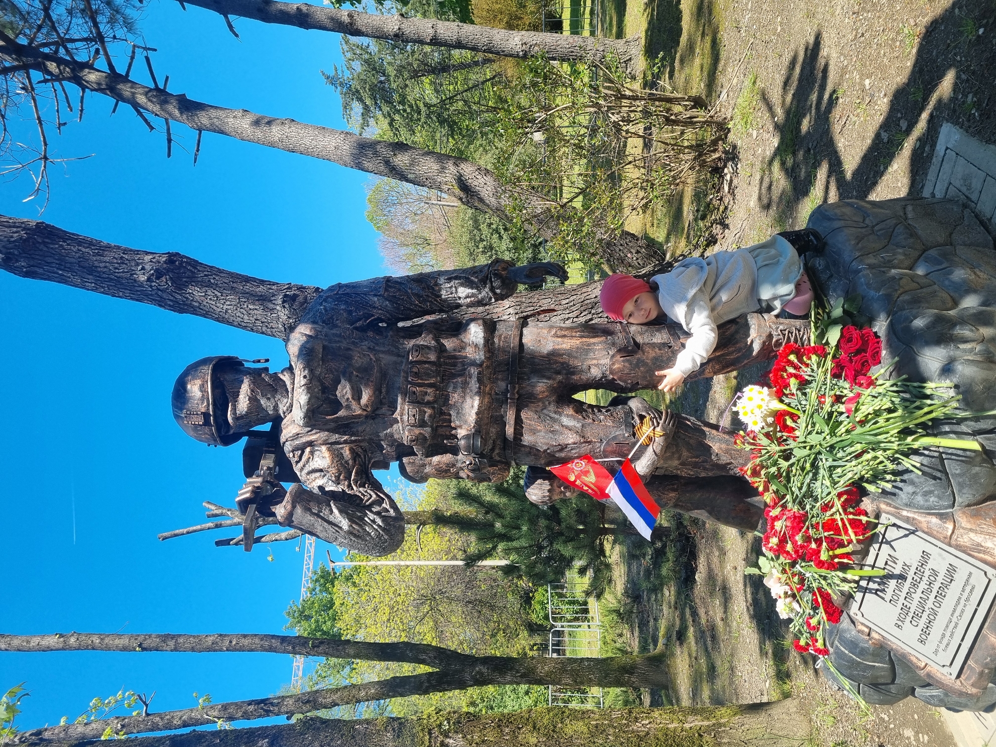 A waist-level shot, rotated 90 degrees clockwise, depicts a statue with a child in front of it. The statue is of a soldier with a rifle, set among trees. A child in a red hat and light-colored clothing is lying on top of a portion of the statue near a bouquet of red and white flowers. To the soldier's left is a Russian flag. A plaque with Cyrillic script sits to the right of the bouquet of flowers. The script mentions the death of soldiers in the "special military operation" and mentions "Sochi" Russia.