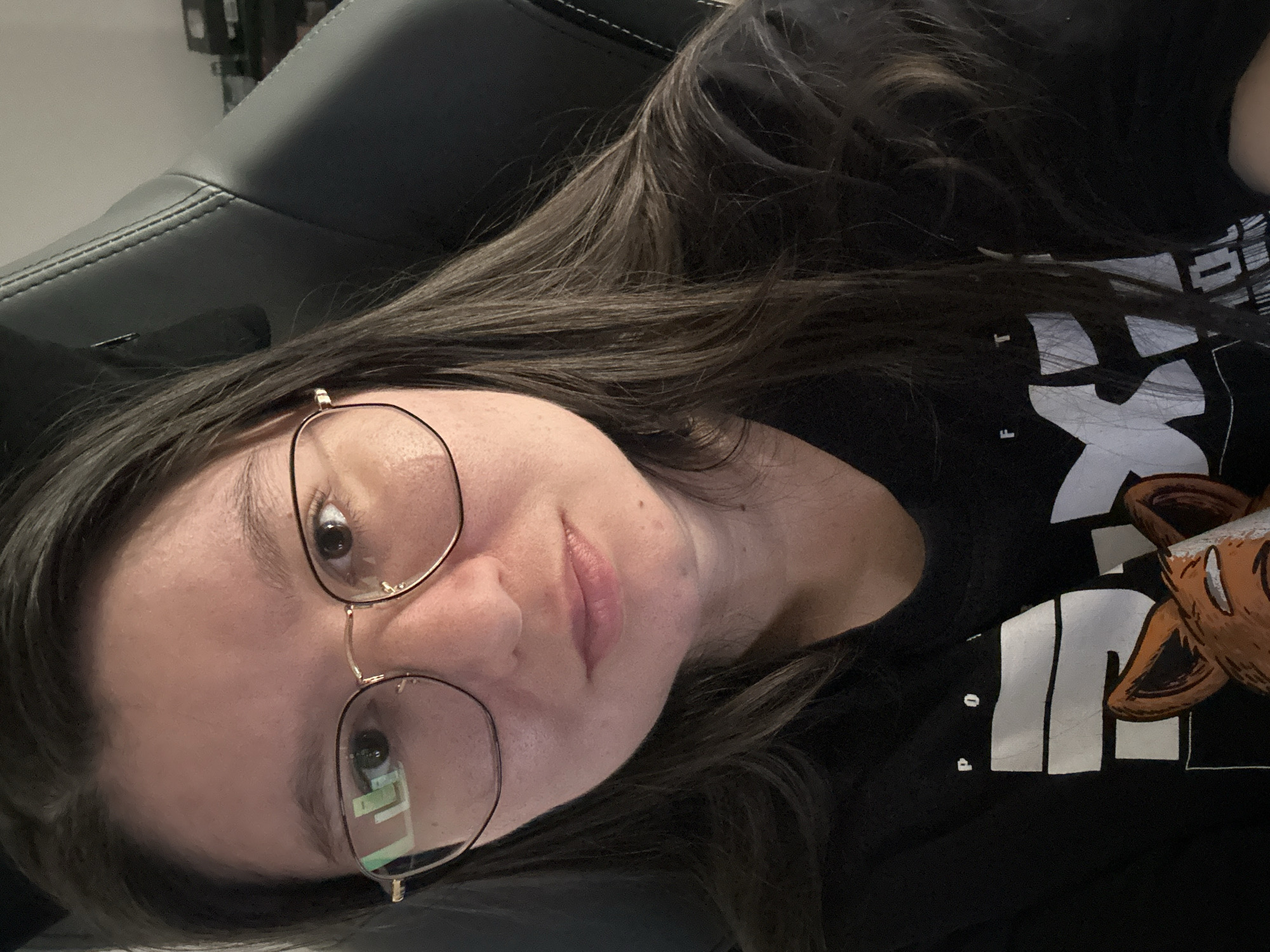 A woman with long, dark brown hair and wearing round, gold-rimmed glasses is looking at the camera. She has fair skin and is wearing a black t-shirt with white lettering and a partially visible brown animal design. She is sitting in a black leather chair. The location in the city of Câmara de Lobos, Portugal, cannot be determined from the image.