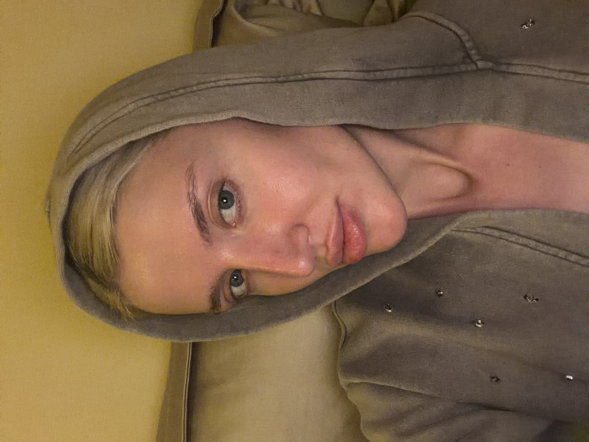 The image features a close-up portrait of a person, likely female, with fair skin and blonde hair, looking directly at the camera with a neutral expression. The person's head is tilted, and their light-colored eyes appear greenish-blue. Their lips are slightly parted.
They are wearing a hooded sweatshirt of a muted khaki-grey or taupe color, with the hood pulled up to frame their face. Small, silvery metallic embellishments, possibly studs or sequins shaped like tiny flowers or stars, are visible on the fabric of the sweatshirt, particularly on the right side of the image. The fabric of the garment shows a soft, textured quality. The person's neck and collarbone are visible, showing natural contours.
The background is indistinct, consisting of a plain yellowish or light beige surface, suggesting an indoor setting, possibly a wall or upholstered furniture. The lighting appears dim and soft. There is no visible text in the image. The scene is set in Nerin, Montenegro.