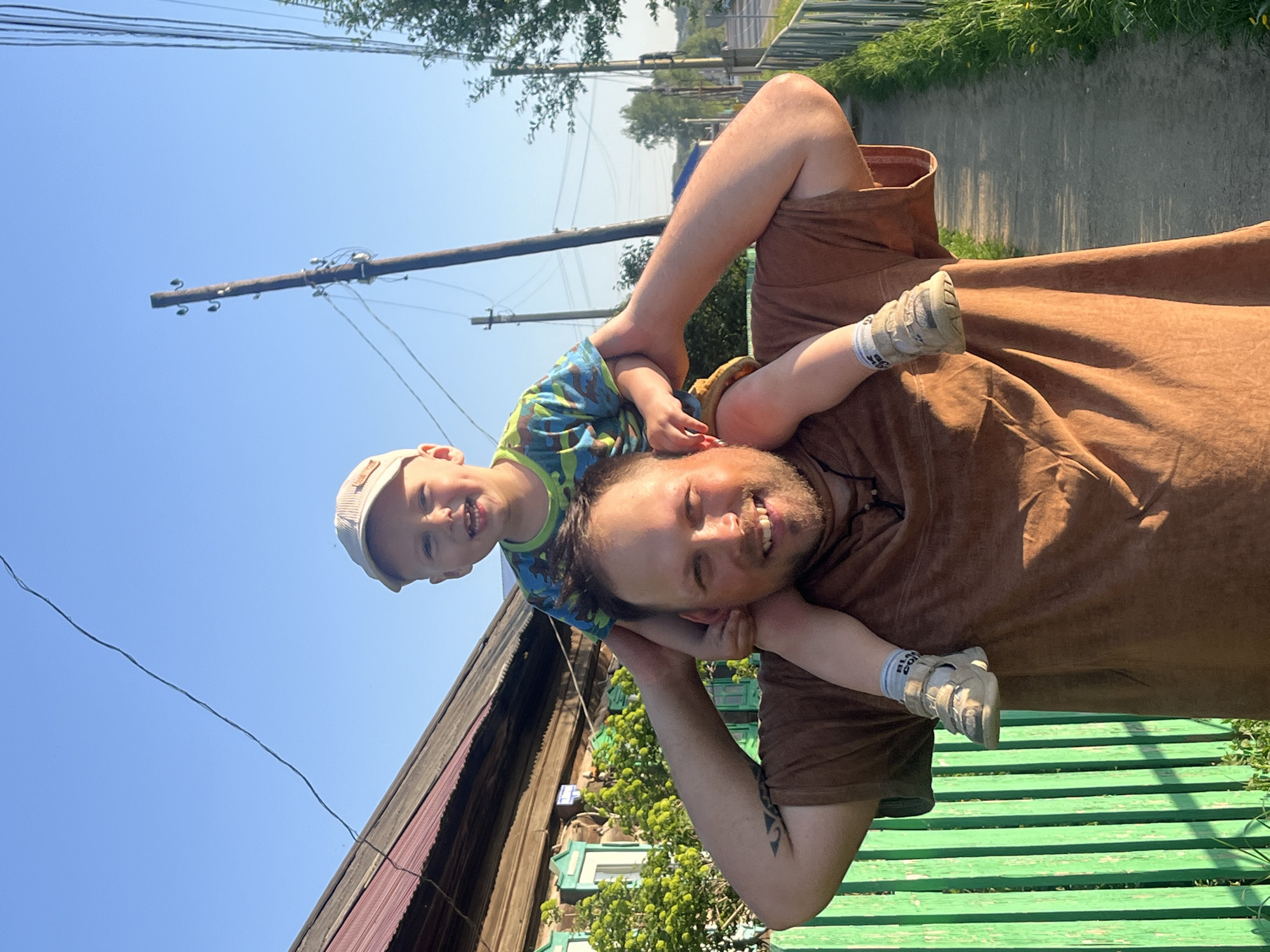 The image shows a man carrying a young child on his shoulders. Both are smiling. The man is wearing a brown shirt and the child is wearing a colorful shirt and a light-colored baseball cap. The child's legs are visible, with small shoes. Behind them is a wooden house with green trim and a corrugated metal roof, along with a green wooden fence. There's a utility pole with wires visible in the background, along with green trees and a path. The location appears to be in the city of Фоки, Russia, judging by the style of the house and general setting.