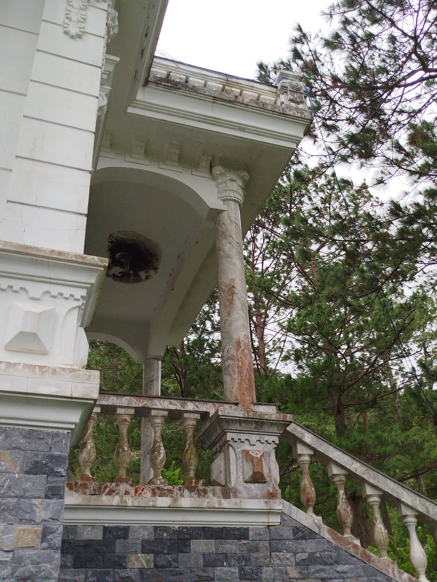 The image shows a portion of an old white building with ornate architectural details. The building features a large column with marble-like veining, a curved archway, and a stone balustrade. In the background, lush green pine trees are visible against a pale sky. The stonework of the building appears weathered and aged, with some cracking and discoloration. This scene is located at the abandoned Domaine de Marie church in Da Lat, Vietnam, a historic landmark known for its French colonial architecture and elevated position offering scenic views of the surrounding landscape.