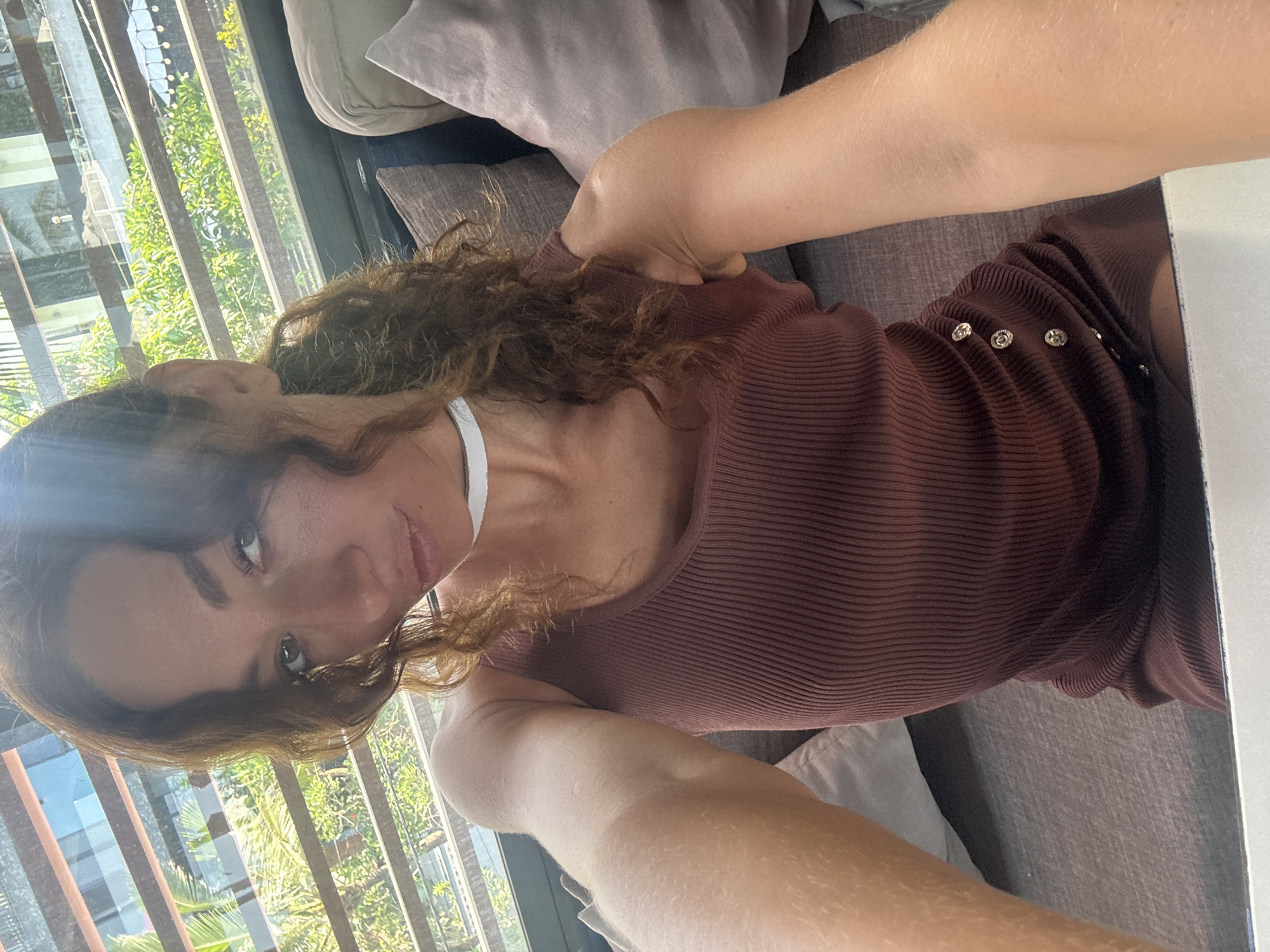 A selfie taken indoors features a woman with curly brown hair, wearing a brown ribbed tank top or dress with decorative buttons on the side. She is positioned on a grey couch with several pillows. Outside the window, foliage and the architectural structure of a building are visible. The scene suggests a casual, relaxed setting in a building with a view of greenery, though the exact location within Đà Nẵng cannot be determined from the visual data.