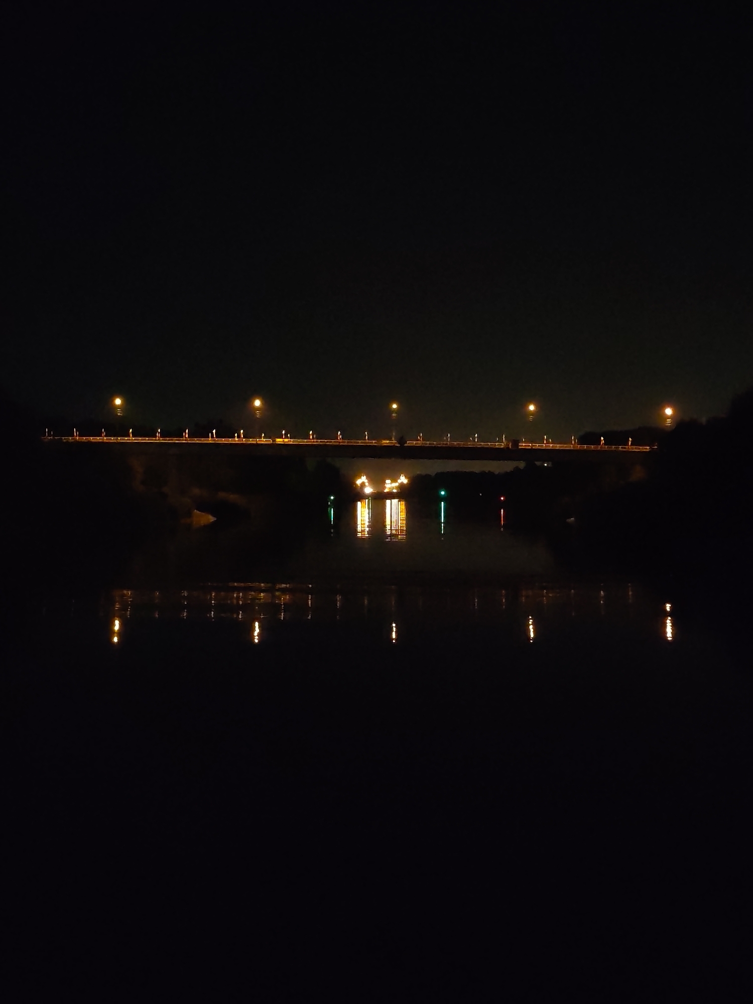 The image depicts a night scene in Yakhroma, Russia, showcasing a bridge illuminated by streetlights, spanning across a body of water. The bridge features a series of evenly spaced lights casting a warm glow onto the structure. Underneath the bridge, lights are visible, potentially from structures or vehicles. Their reflections shimmer on the water's surface, creating vertical streaks of light. The water is surrounded by dark, undefined shapes suggesting land covered with vegetation or structures. The sky is a dark, featureless void, emphasizing the contrast between the artificial lights and the natural darkness. No people or specific actions are discernible in the image.
