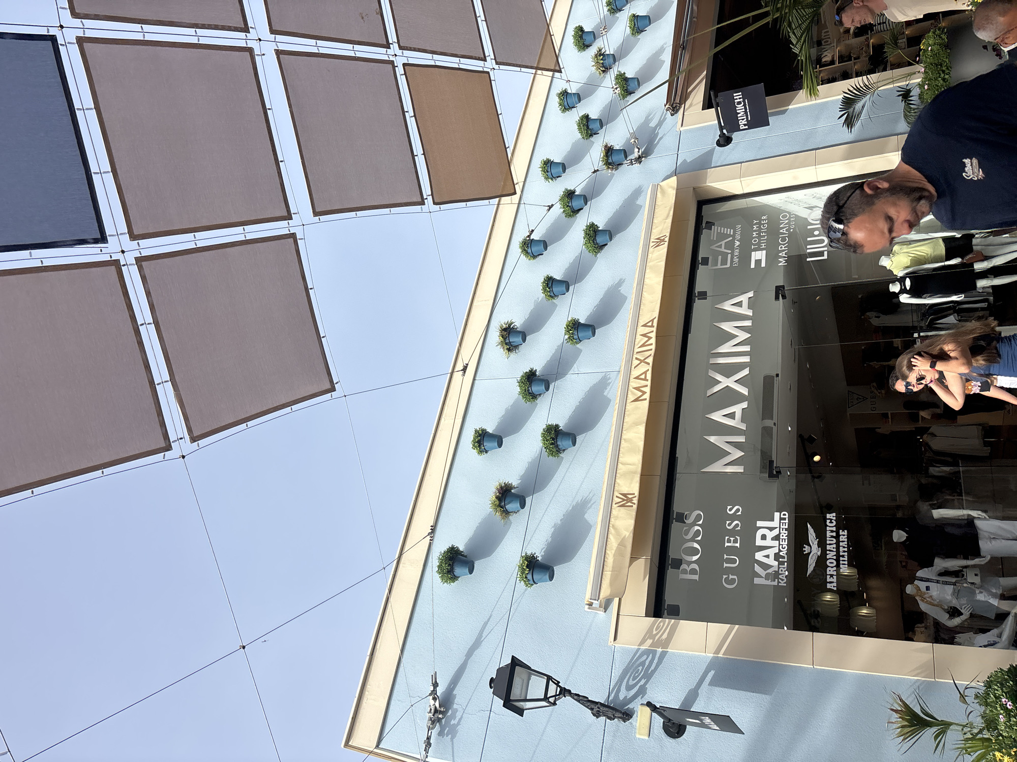 The image shows the exterior of a store named "MAXIMA" in Orihuela, Spain. The store's facade is light blue with a beige awning above the entrance. The entrance is a large glass storefront displaying various brand names such as BOSS, GUESS, KARL LAGERFELD, AERONAUTICA MILITARE, EA7 EMPORIO ARMANI, TOMMY HILFIGER, MARCIANO, and LIU-JO. Inside, mannequins are visible displaying clothing.
Above the store, a series of square, brown and dark fabric shades are suspended by wires against a clear blue sky, providing shade. The light blue wall of the building is adorned with numerous small, blue potted plants arranged in a grid pattern. A black vintage-style street lamp is mounted on the left side of the building, casting a shadow.
In the foreground, a man with short dark hair, wearing sunglasses on his head and a dark t-shirt, is walking towards the right of the frame, looking downwards. To his left, a woman with long brown hair and sunglasses is also visible. Another man is partially visible on the far right of the image. The ground area is paved.