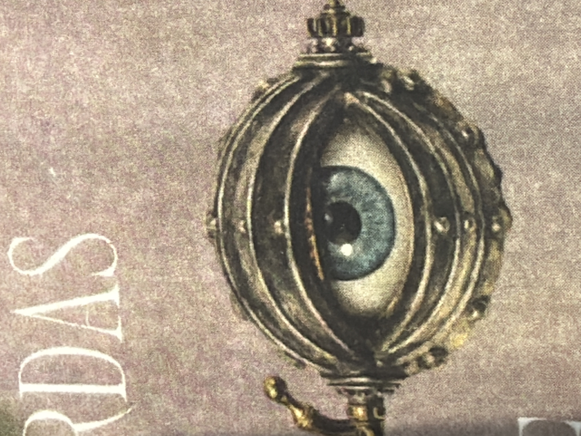 The image displays a close-up of an intricate illustration featuring a mechanical eye against a mottled, textured reddish-brown background. The eye itself exhibits a vivid blue iris with a central black pupil and a white sclera, framed by a complex, multi-layered metallic housing. This housing appears to be constructed from aged or tarnished metal, possibly bronze or darkened brass, and is detailed with rivets and segmented panels. An ornate, golden-hued lever or handle projects from the left side of the mechanical casing, while a small gear mechanism is visible on the right. Partially visible white sans-serif letters "RDAS" are positioned across the top of the image. The scene is devoid of people or discernible actions. No specific visual elements or landmarks within the image identify a location in the city of San Andrés Cholula, Mexico.