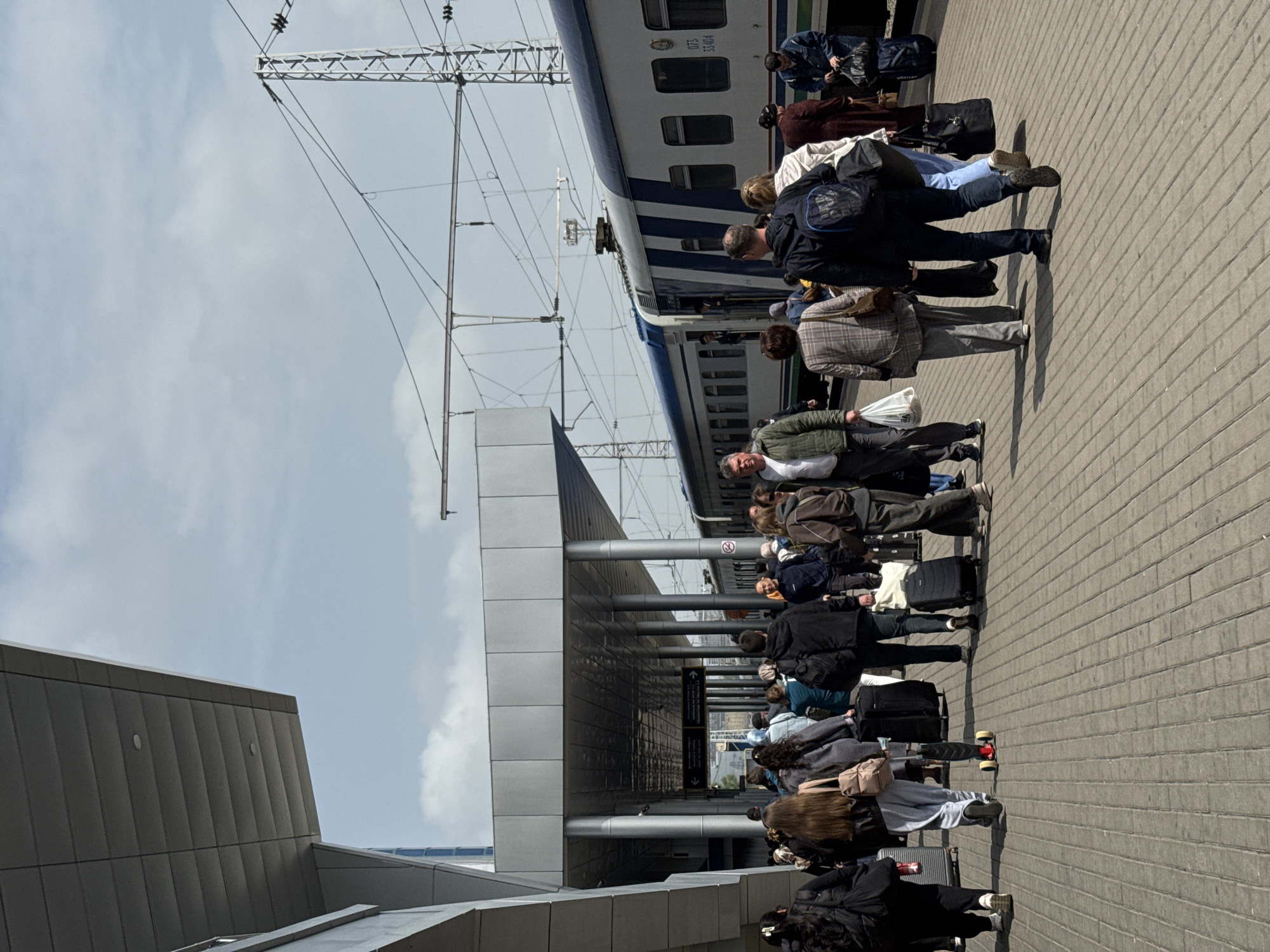 People are lining up at the Tashkent train station in Uzbekistan to board a train. They are standing on a paved platform next to the train tracks. Most of them have luggage with them. The train is blue and white. Overhead power lines are visible above the train. The sky is partly cloudy.