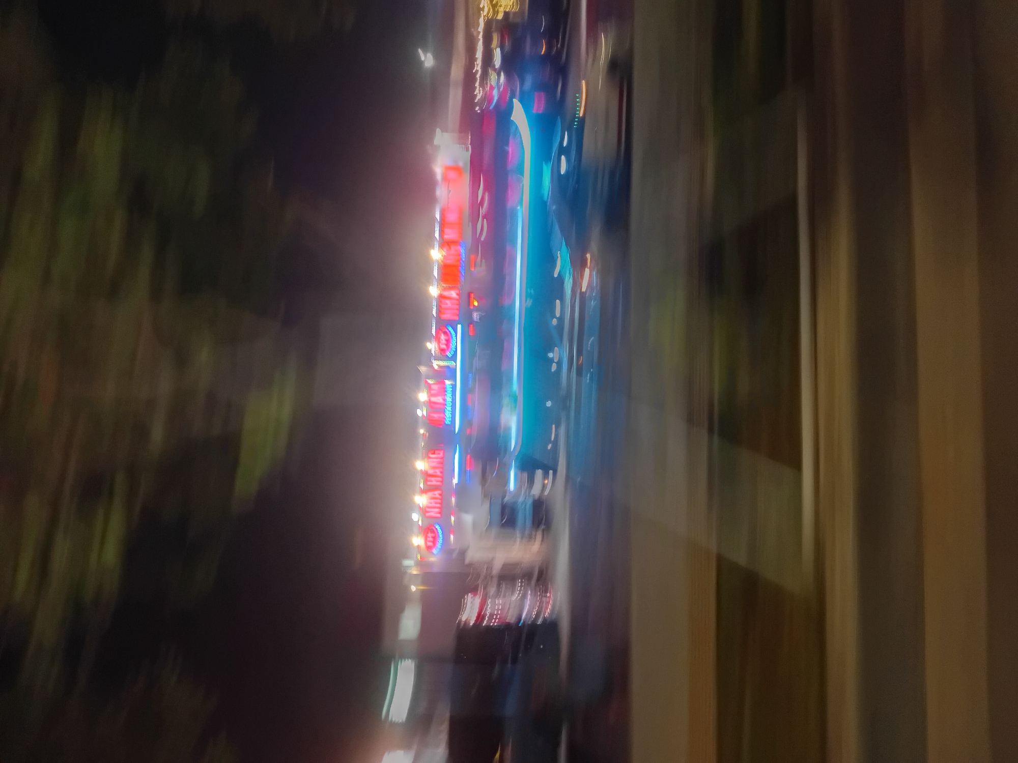 The image is a motion blur of a street scene at night in Da Nang, Vietnam. A brightly lit restaurant sign reads "NHÀ HÀNG", indicating a dining establishment. Several vehicles, possibly including trucks and cars, are visible, their lights streaked due to the movement. The overall impression is of a dynamic, urban environment with a focus on commercial activity and transportation at night. The foliage of trees is visible in the background, suggesting a street lined with greenery. The foreground shows a road surface and possibly a sidewalk or median strip, also blurred by motion.