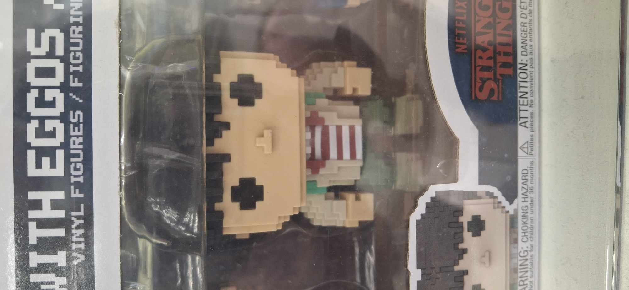 The image shows a close-up view of a Funko Pop vinyl figure in its packaging. The figure is pixelated in design and depicts a character from "Stranger Things" wearing a red and white striped shirt and a black hat. The packaging displays the "Netflix" logo and "Stranger Things" title. Below the figure, there is a warning label in English and French indicating a choking hazard and that the product is not suitable for children under 36 months. The background is blurred but appears to be an indoor setting, possibly a retail store. The lighting is artificial, and there is some glare on the plastic packaging. The image does not provide any information about the location being Saint Petersburg, Russia.