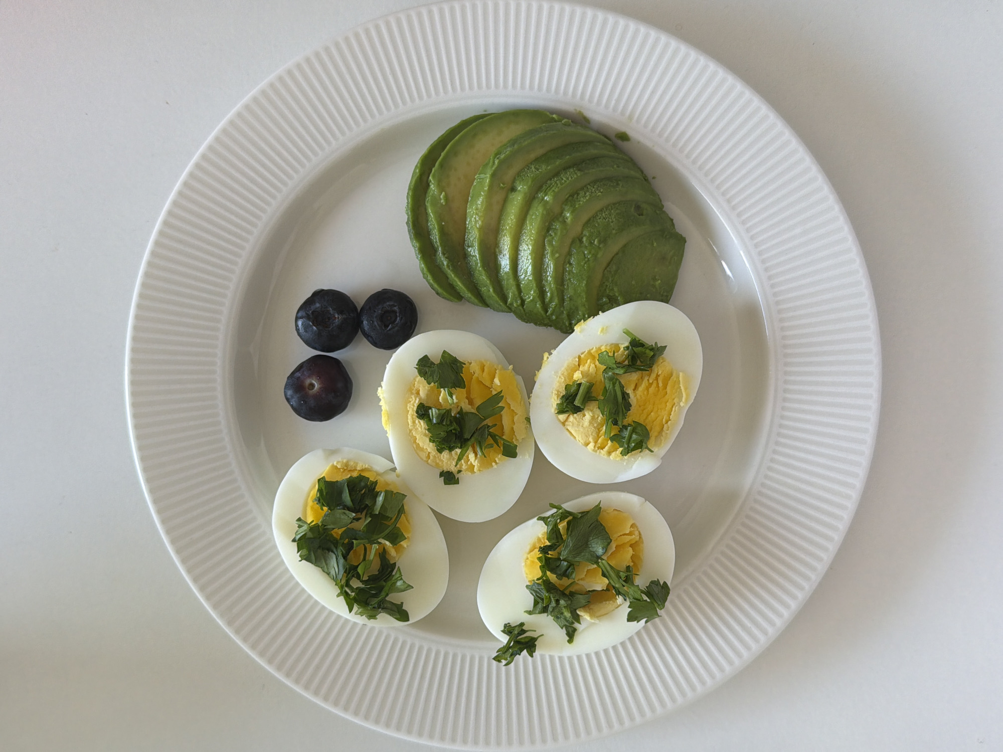 A top-down view reveals a white, circular plate holding a small assortment of food, consisting of sliced avocado, four halves of hard-boiled eggs sprinkled with green herbs, and three blueberries. The plate sits on a plain white surface. The image does not provide information about the location in Tel Aviv or any people, actions, or interactions.
