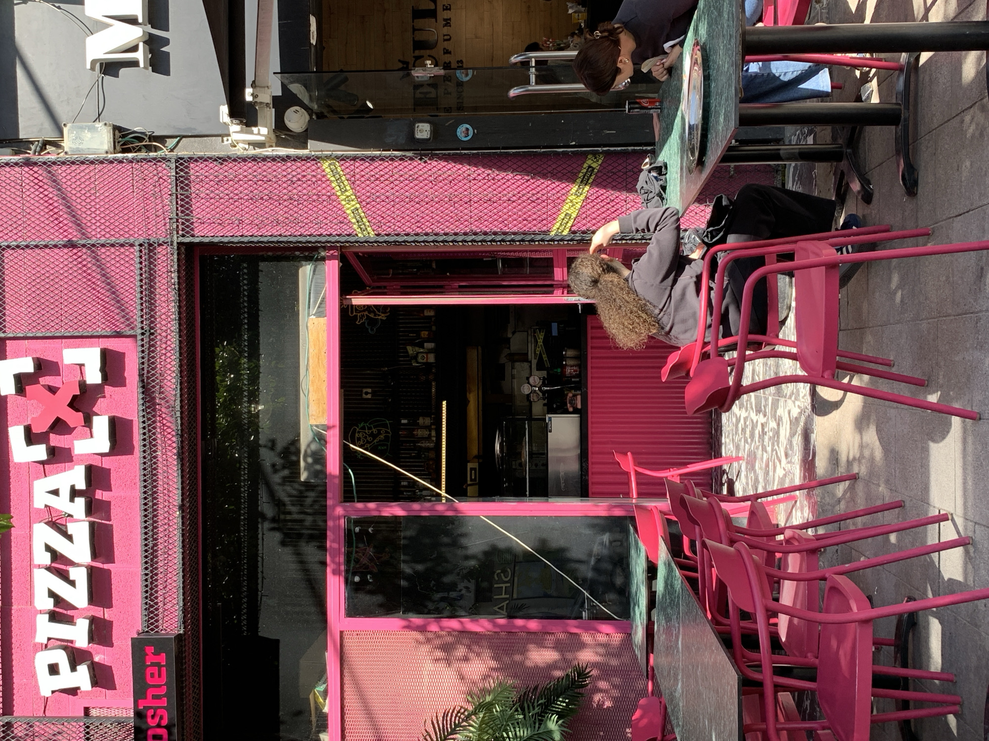 The image captures a street scene in Tel-Aviv, Israel, featuring a pink-themed pizzeria with the word "PIZZA" displayed vertically in white letters on a sign. The pizzeria is decorated with pink paint and mesh accents, and the word "Kosher" is visible on the signage.
Two women are present. One woman is seated at a table, seen in profile from the shoulders up; the second woman is seated on one of the pink chairs in front of the pizzeria. The woman is wearing dark clothing and has long, curly hair.
A row of pink metal chairs and tables is lined up along the sidewalk in front of the pizzeria. The chairs are stacked on top of each other, suggesting they are not currently in use.
The scene appears to be in daylight with shadows cast on the building's exterior and the sidewalk. The overall impression is a colorful, urban setting with a food establishment as the central focus.