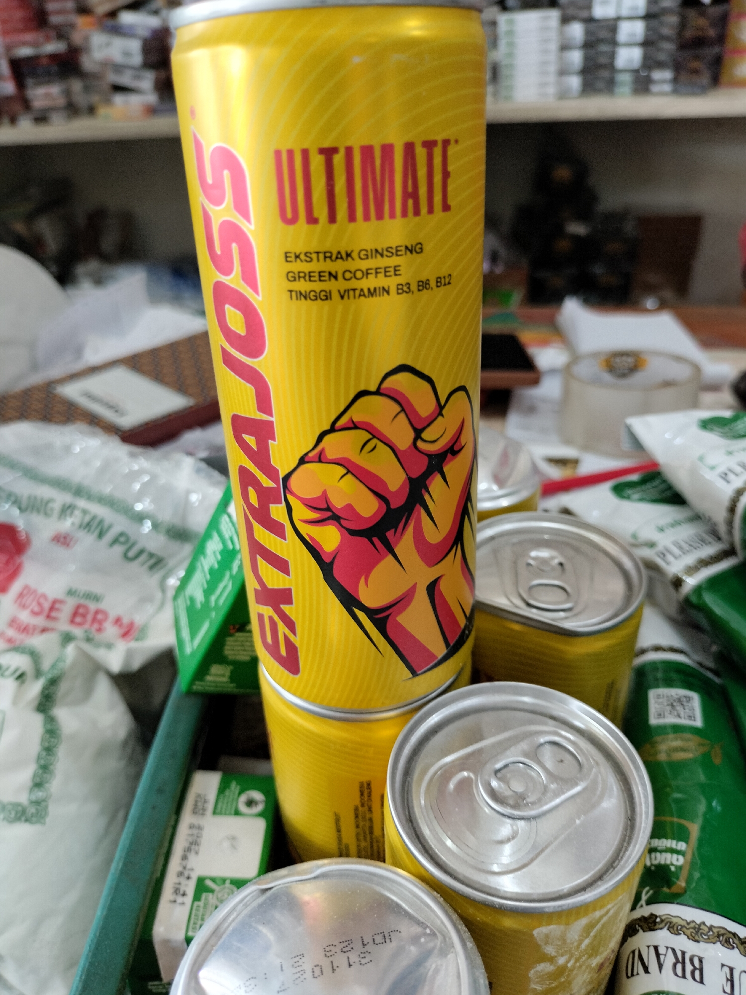 In Meudangara, Indonesia, a close-up shot focuses on a collection of beverage cans, primarily energy drinks, arranged on a surface. A prominent yellow can with red lettering, labeled "EXTRA JOSS ULTIMATE," stands out. The can features text indicating "EKSTRAK GINSENG," "GREEN COFFEE," and "TINGGI VITAMIN B3, B6, B12." An illustration of a clenched fist is visible on the can. Several other yellow and silver cans are partially visible, some stacked. In the background, shelves are stocked with various items, appearing to be a retail or storage environment. The overall scene suggests a commercial setting.