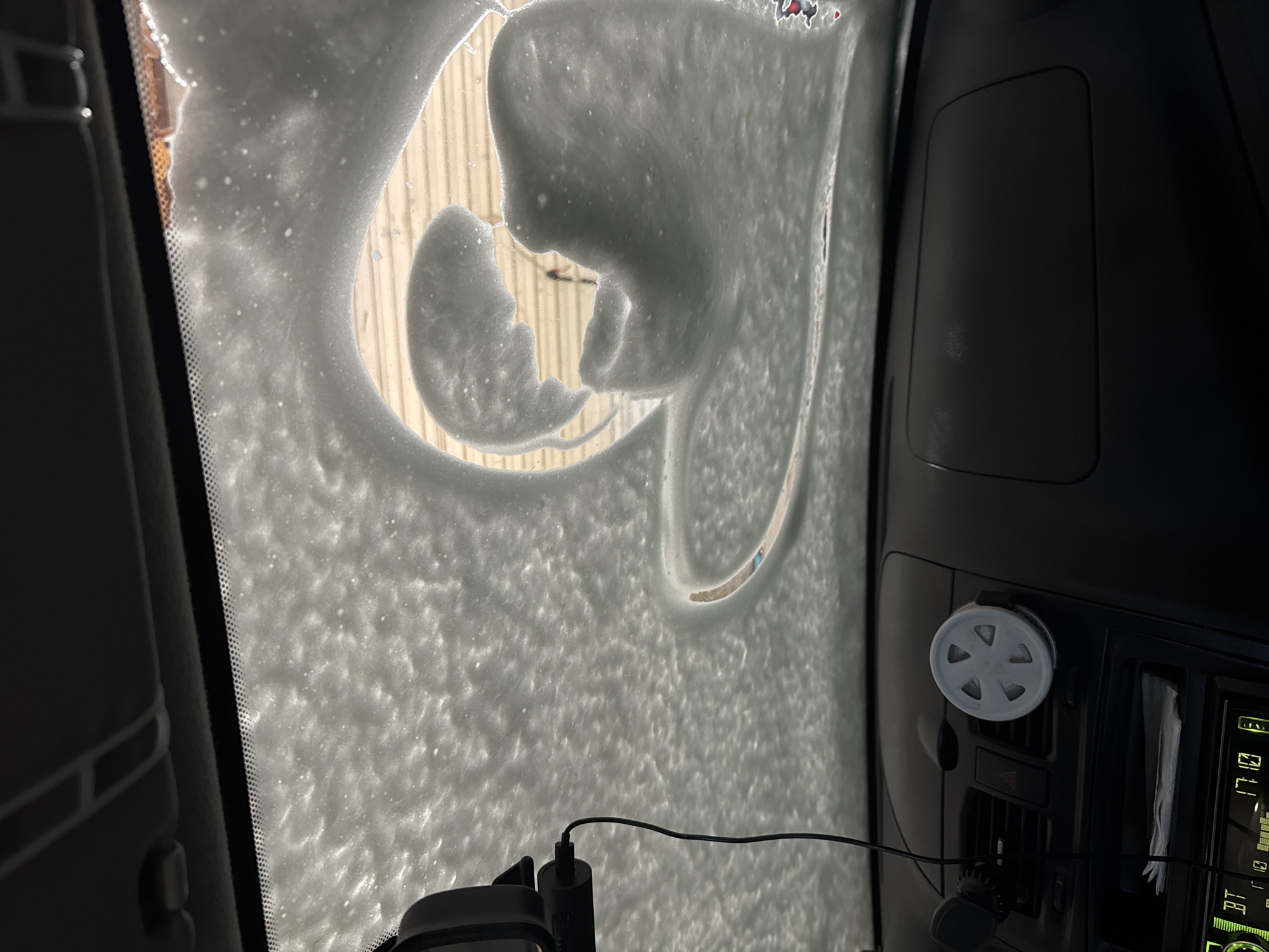 View from inside a vehicle, likely a car, through the windshield. The windshield is heavily coated with white foam, resembling car wash soap or snow. Patterns in the foam suggest that it has been sprayed or applied in a non-uniform manner. Outside, visible through a few partially cleared sections of the windshield, is what appears to be an industrial-type building with corrugated metal siding. The interior of the vehicle shows part of the dashboard, including an air vent with a white dial, a wire cable, and an electronic device with a screen displaying numbers and letters. The scene provides no indication of its location within Perm, Russia.