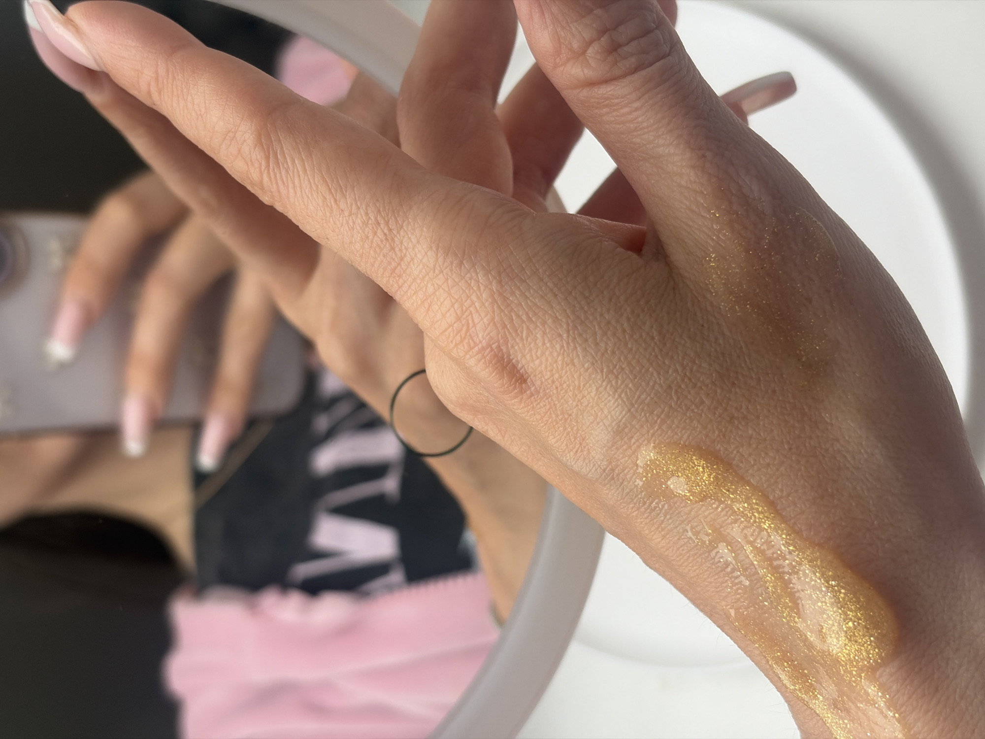 The image shows a close-up of a person's hand with a gold, glittery liquid on the back of the hand, presumably a lotion or similar cosmetic product. The person is holding their hand in front of a mirror. The reflection shows a blurred image of the person holding a phone and wearing a pink top. The background includes a partial view of a white surface. There is no discernible information to indicate that this image was taken in Novosaratovka, Russia.