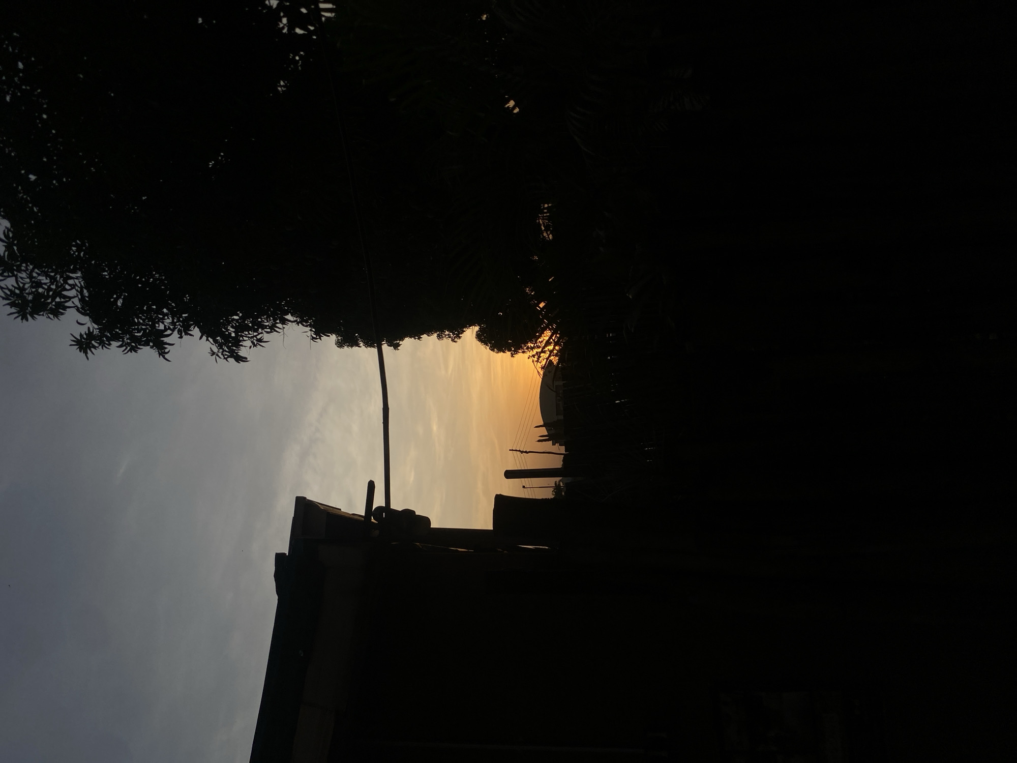 The image depicts a serene sunset or sunrise scene in Barden Yana, Nigeria, characterized by dramatic silhouettes against a gradient sky. The sky transitions from a cool, light grey on the left to a warm, glowing orange-yellow near the right horizon, indicating either dawn or dusk. Wispy clouds are scattered across the cooler parts of the sky.
In the foreground, the dense foliage of trees forms a dark, intricate silhouette along the top and bottom left sections of the frame. On the bottom right, the sharp, angular silhouette of a building's roofline is prominently visible, topped with a few antenna-like structures. Against the vibrant orange sky in the midground, several utility poles with power lines create a series of vertical and horizontal dark lines. Further in the distance, the faint silhouettes of other structures or buildings can be seen.
The photograph is dominated by strong contrasts between the bright, colorful sky and the almost completely black, underexposed foreground and midground elements. This creates a peaceful and contemplative mood, highlighting the natural beauty of the light and shadow. No specific activity or individuals are depicted, and there is no visible text within the image.