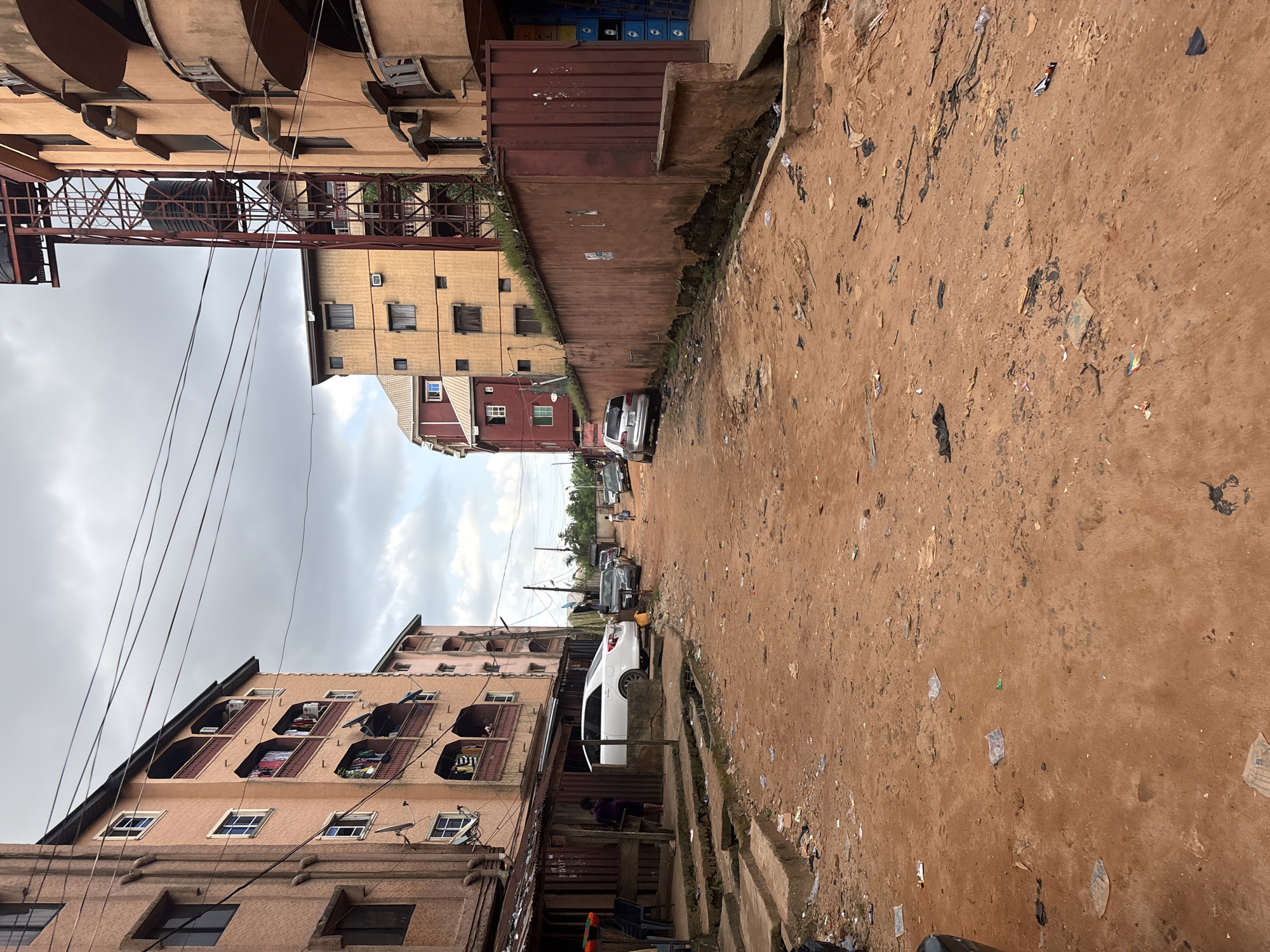The image depicts a street scene in Onitsha, Nigeria, characterized by a dusty, reddish-brown dirt road. On either side of the road are multi-story buildings, appearing to be apartment complexes. The architecture suggests a dense urban environment. The buildings on the left have balconies with laundry hanging out to dry, indicating residential use. A white sedan is parked near a small, weathered structure on the left side of the road, partially obscuring a set of concrete steps leading up to the building. Several other cars are visible further down the road, parked along the sides.
The road itself is unpaved and shows signs of wear and tear, with visible tire tracks. Scattered across the surface are pieces of debris, including plastic bags, paper, and other refuse, suggesting a lack of consistent waste management. The overall impression is one of a bustling, perhaps somewhat impoverished, urban area. The sky is overcast, contributing to a muted lighting condition. A person, a woman, is visible standing near the structure on the left, and a child is seen walking in the distance on the right side of the road. The scene is comprised of both residential structures and evidence of everyday life in a busy city.
