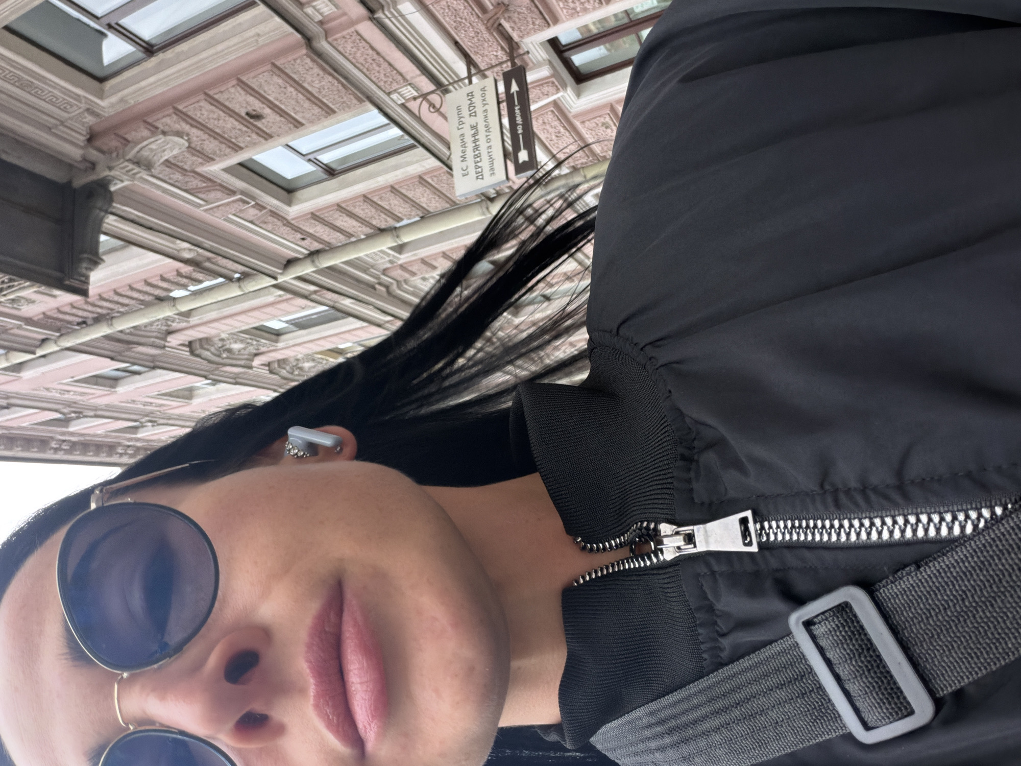 A person is seen from a low-angle perspective in what appears to be Saint Petersburg, Russia. The person is wearing sunglasses and an earbud, and a black jacket. A sign in Russian is visible in the background, reading "ЕС Медиа Групп ДЕРЕВЯННЫЕ ДОМА защита отделки уход" and an arrow pointing towards "ВО ДВОРЕ". The architecture suggests an urban setting.