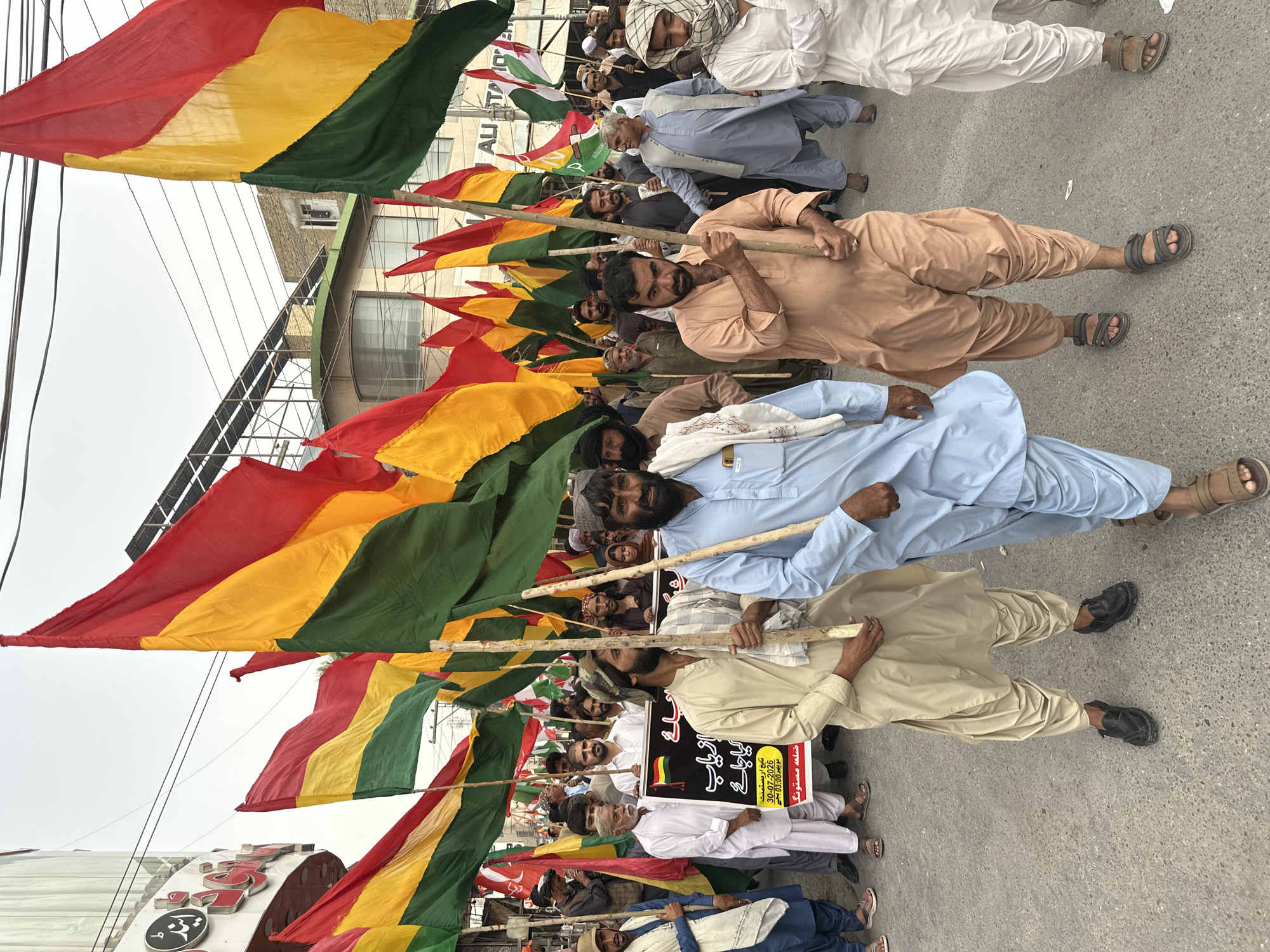 A group of men are participating in a street protest in Quetta City Tehsil, Pakistan. They are holding up multiple flags, predominantly in red, yellow, and green horizontal stripes. Some of the men are wearing traditional Pakistani attire, such as shalwar kameez, while others are in more casual clothing. Many of the men are holding wooden poles that support the flags. In the background, there are buildings and overhead power lines, indicating an urban setting. One man in the foreground is holding a banner with text in Urdu. The overall atmosphere appears to be a public demonstration.