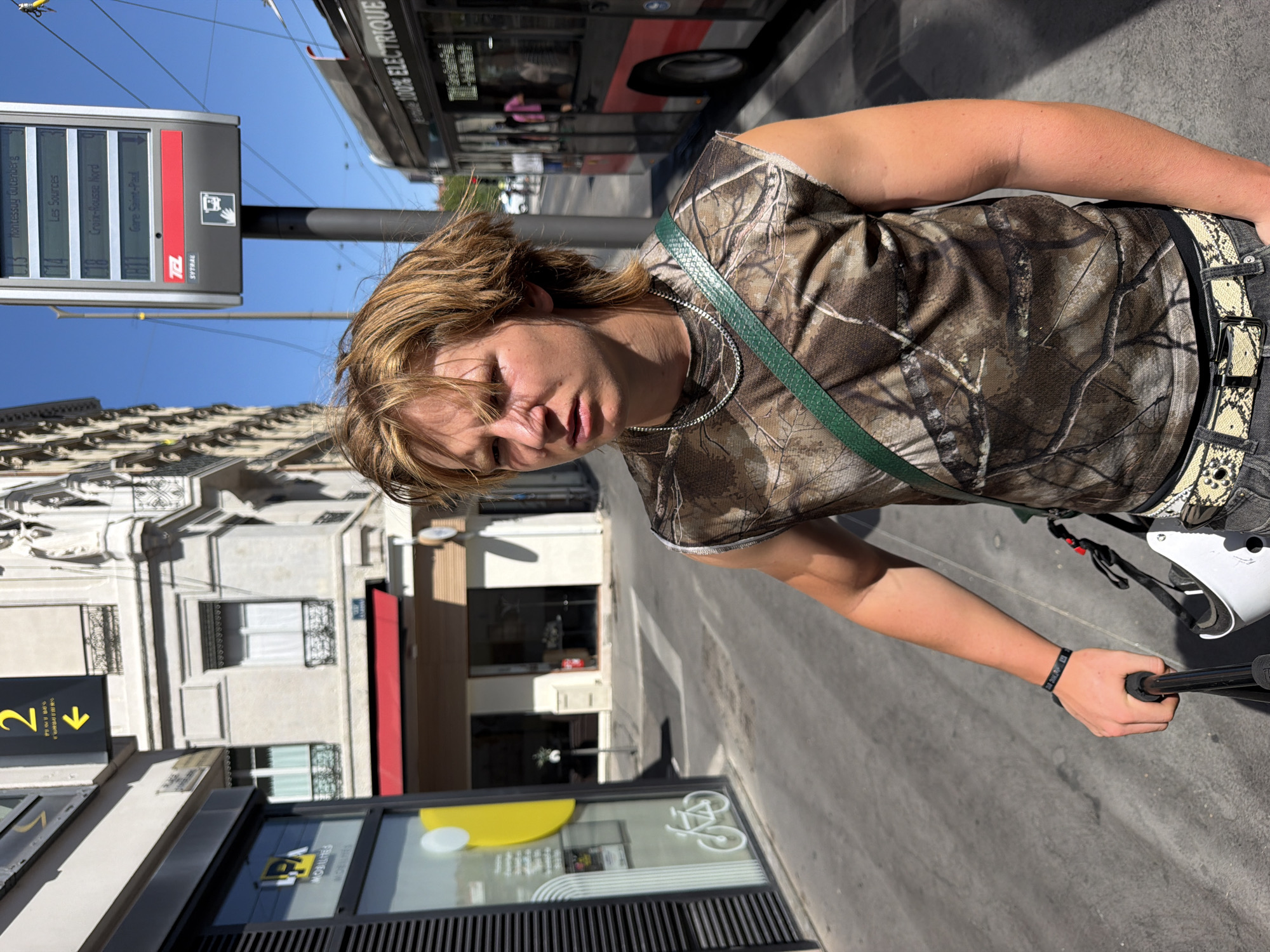 A young person with blonde, shaggy hair stands on a sidewalk in Lyon, France, holding a selfie stick. The person is wearing a sleeveless camouflage shirt, a green strap across their chest, and a snakeskin-patterned belt over jeans. Behind them, a TCL (Transport en Commun de la Agglomération Lyonnaise) bus and a bus stop information display are visible. The bus stop display shows route information for TCL, including stops like "Montessuy Gutenberg" and "Croix-Rousse Nord." Buildings with classical architecture line the street, and a bright blue sky indicates it is daytime. The scene appears to be a public street in an urban environment.