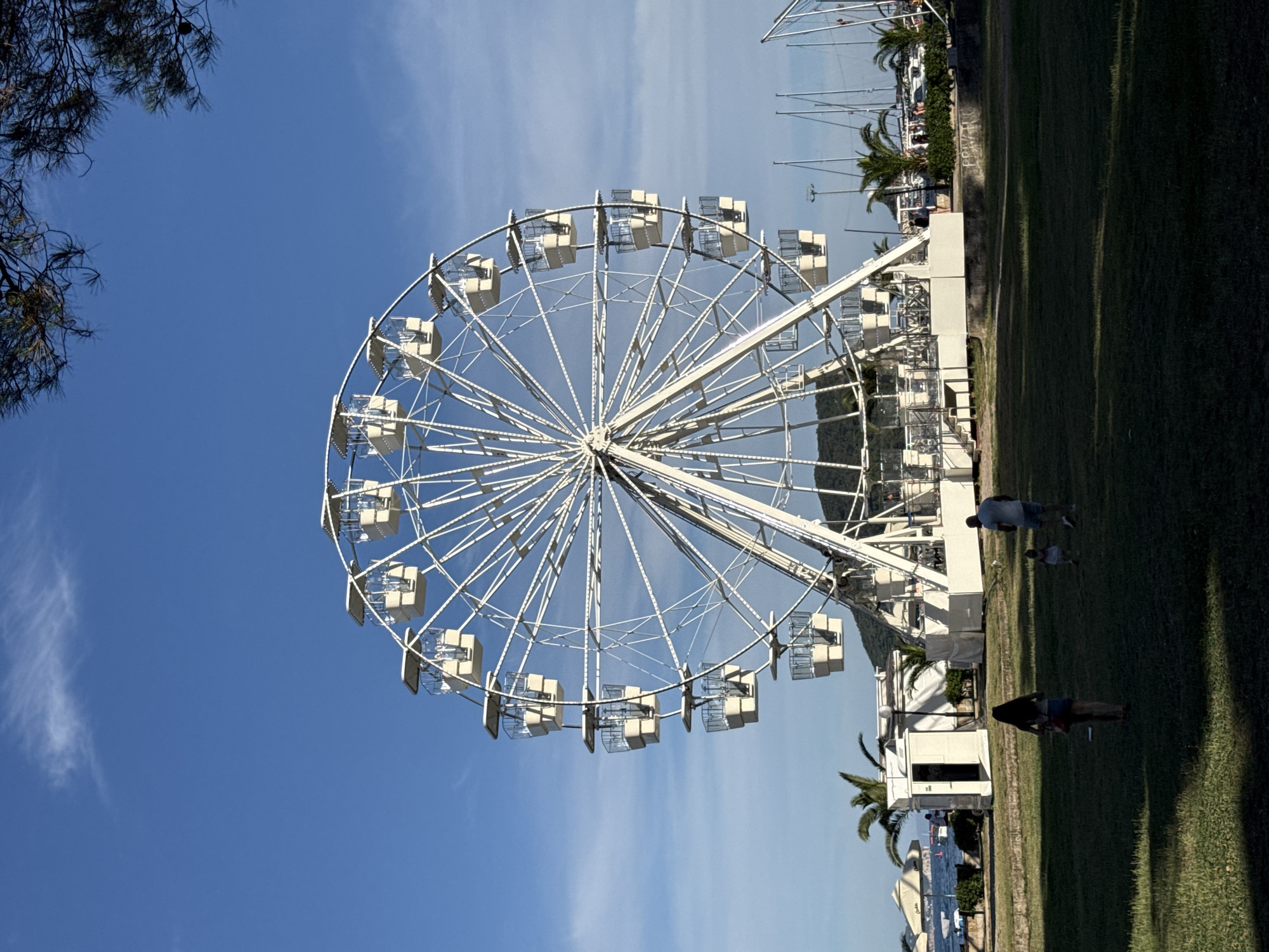 A white Ferris wheel is centrally located in the frame, with its structure dominating the upper portion of the image. The Ferris wheel has multiple enclosed cabins, each with clear windows. The sky above is a vibrant blue with a few wispy clouds. In the foreground, a grassy area is visible, with shadows cast across it. Three figures are present on the grass: a woman with long dark hair walking away from the camera, a small child running ahead of her, and a man walking in the same direction as the woman, slightly behind her. To the left of the Ferris wheel, a white building with an open door is situated, with palm trees and beach umbrellas visible beyond it near the water. In the background, the sea and a distant hilly landscape can be seen. The scene appears to be in a coastal city, likely Budva, Montenegro, given the presence of palm trees, a marina with sailboats, and a seaside promenade. The overall atmosphere suggests a pleasant, sunny day at a resort or amusement area.