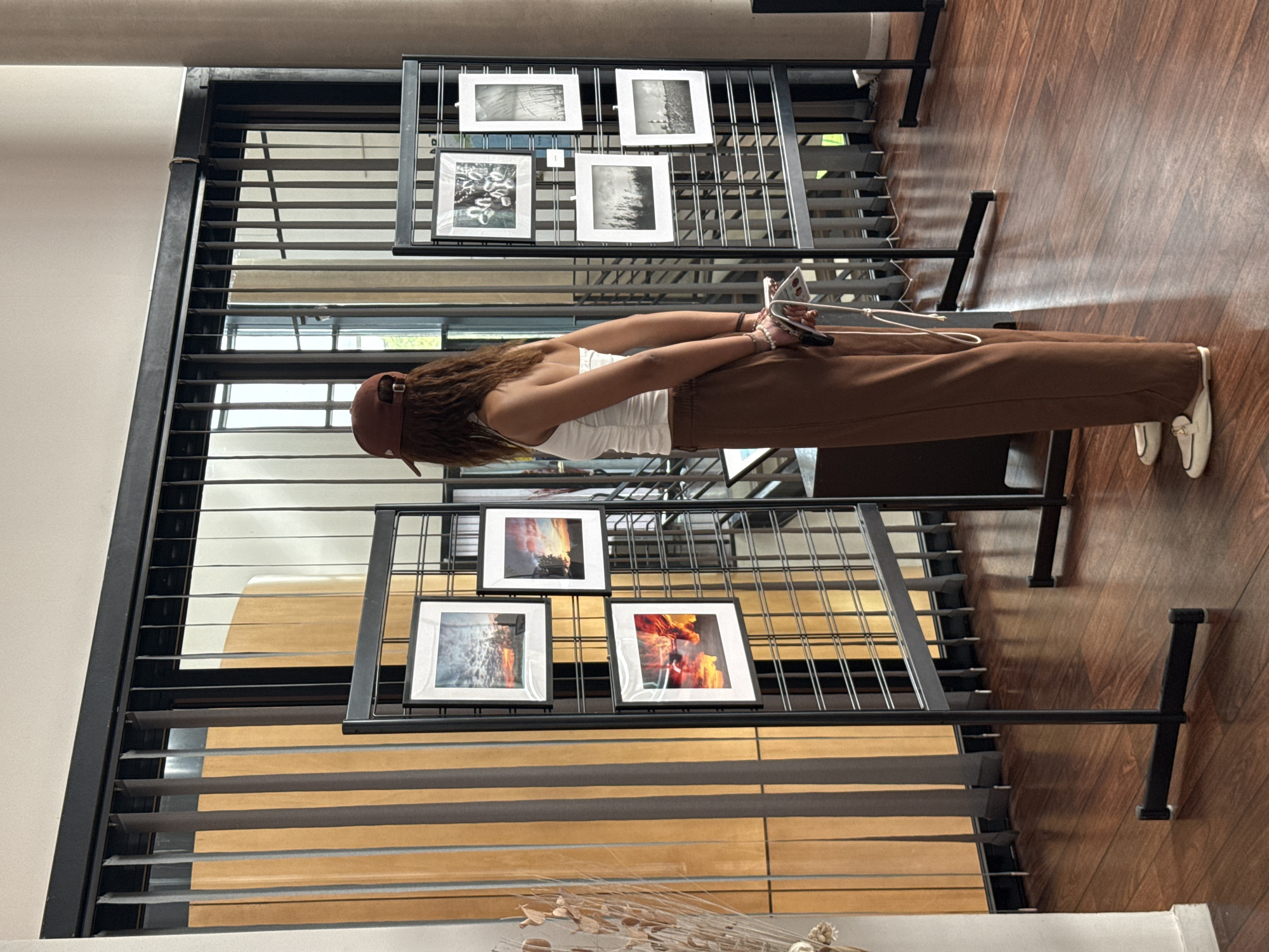 A woman with brown hair and a brown baseball cap, wearing a white tank top and brown pants with white shoes, stands in front of a metal grid display of framed photographs. The photographs are arranged in two tiers and depict various subjects, including landscapes and abstract scenes. The display is situated on a wooden floor. Behind the display, there is a wall with horizontal blinds. The scene is illuminated with natural light. While the location is in the city of Domont, France, there is no information given in the image to confirm this.