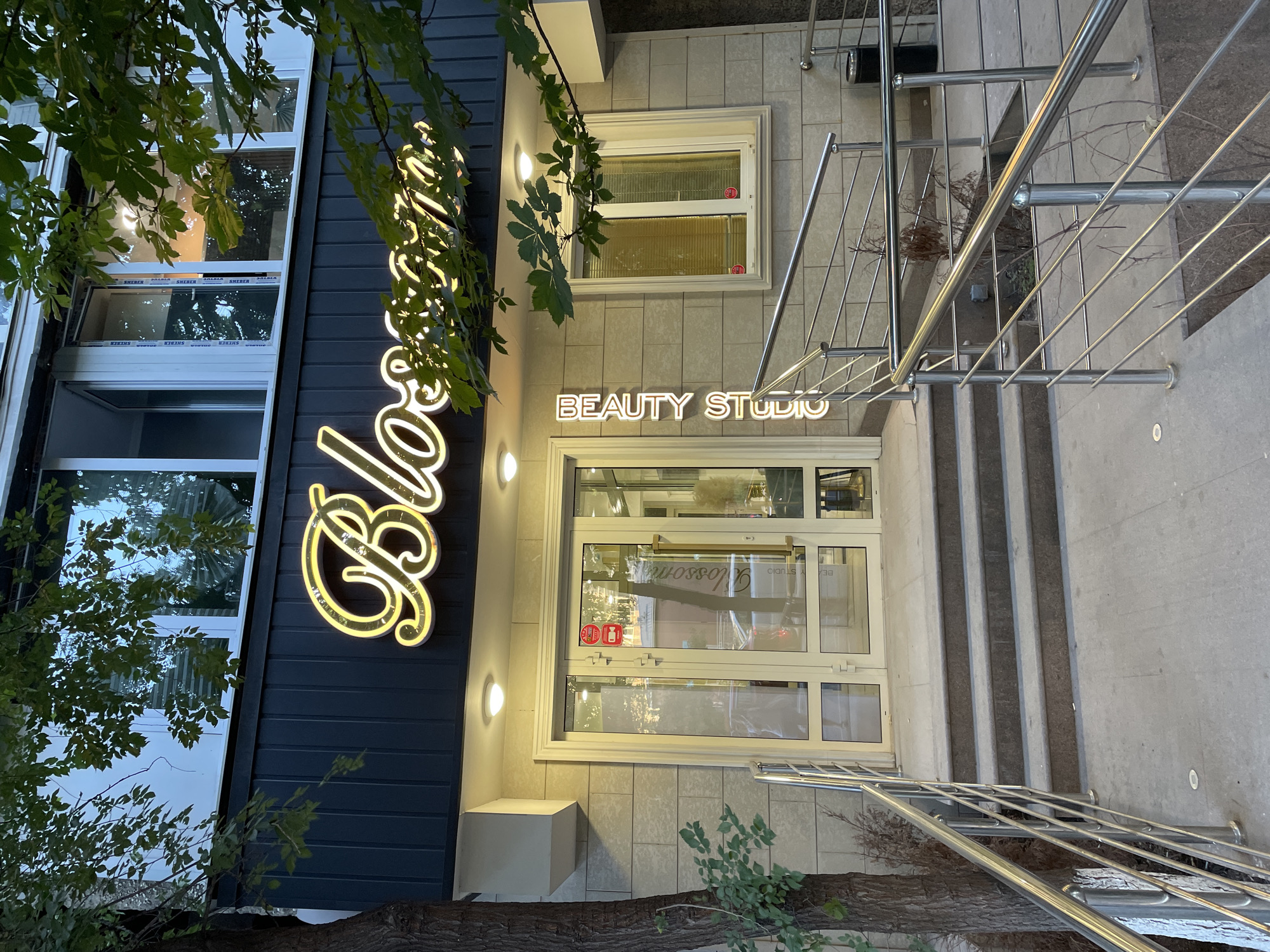 The image shows the exterior of a "Blossom Beauty Studio" in Almaty, Kazakhstan. The studio features a prominent neon sign with the name "Blossom" in cursive script, glowing yellow against a dark facade. Below the sign, a glass-paned entrance door is visible, flanked by stone-tiled walls. A vertical neon sign on the left side of the entrance reads "BEAUTY STUDIO". A set of stone steps leads up to the entrance, with polished metal handrails on either side. Green foliage from trees overhangs the building, partially obscuring the upper windows, which have blinds. The scene appears to be captured during daylight hours, with some artificial lighting visible around the entrance.