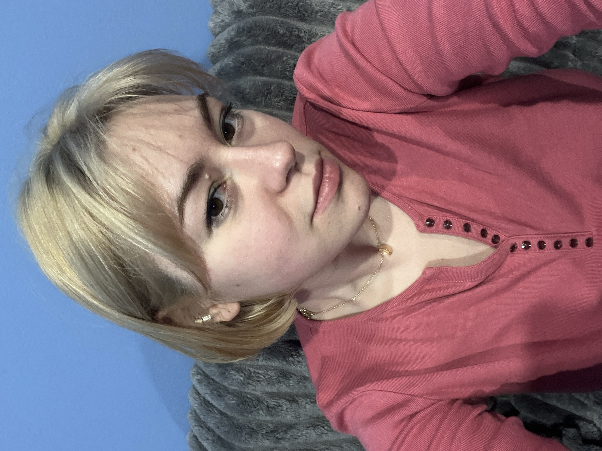 This is a portrait-style, slightly tilted selfie of a young woman taken indoors in Odesa, Ukraine. The woman, who appears to be in her late teens or early twenties, has fair skin, brown eyes, and short, light blonde hair. She looks directly at the camera with a calm, neutral, and slightly contemplative expression, her lips softly parted.
She is wearing a long-sleeved top in a dusty rose or pink hue, which features a row of small, dark, possibly studded or button-like embellishments along the neckline. A delicate gold-toned chain necklace with a small crescent moon pendant is visible around her neck, and a simple, small earring adorns her left ear.
The woman appears to be reclining or lying down, with her head resting on a textured, grey, possibly faux-fur or fluffy blanket or pillow. A plain, solid blue wall serves as the backdrop behind her. The lighting is soft and even, indicating an indoor setting without direct harsh light. There is no visible text in the image.