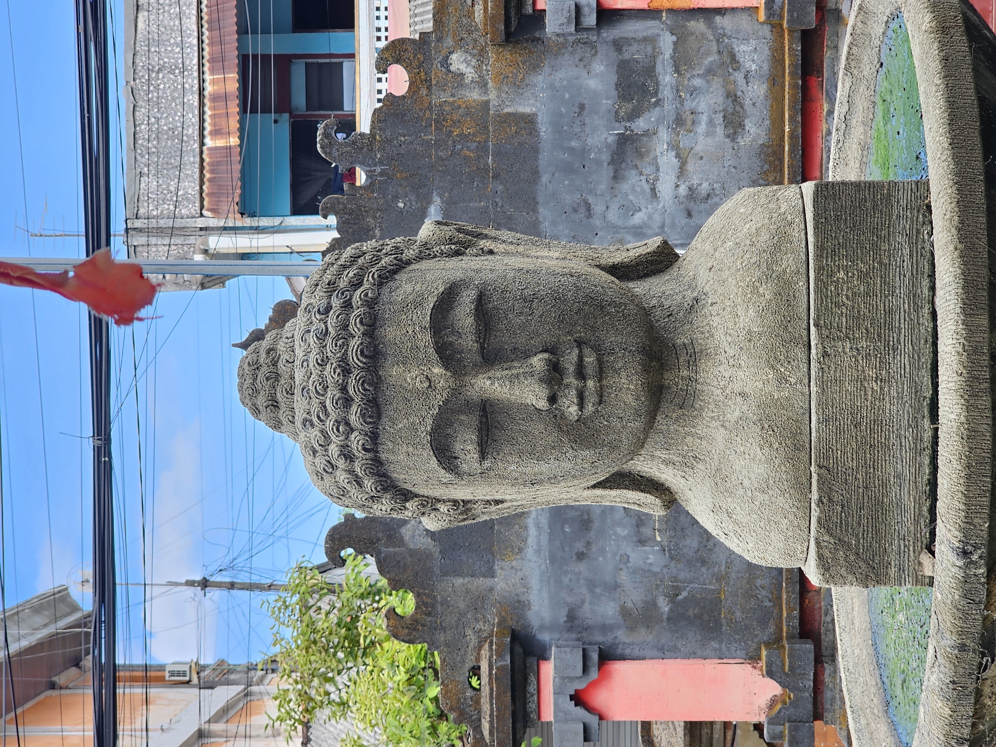 This media file shows a stone statue of Buddha's head and upper body in what appears to be an outdoor setting. The statue is carved with intricate details in the hair and facial features, with eyes closed in a meditative pose. It's positioned in front of a dark stone wall with decorative architectural elements. To the left of the statue, there are buildings with corrugated metal roofs and visible electrical wires against a clear blue sky. Some green foliage is also visible in the lower left and right of the frame. The overall scene suggests a cultural or religious site, possibly in Denpasar, Indonesia, given the context. The lighting indicates it is daytime, with bright sunlight. No specific activity or event is depicted, and no text is visible in the image.