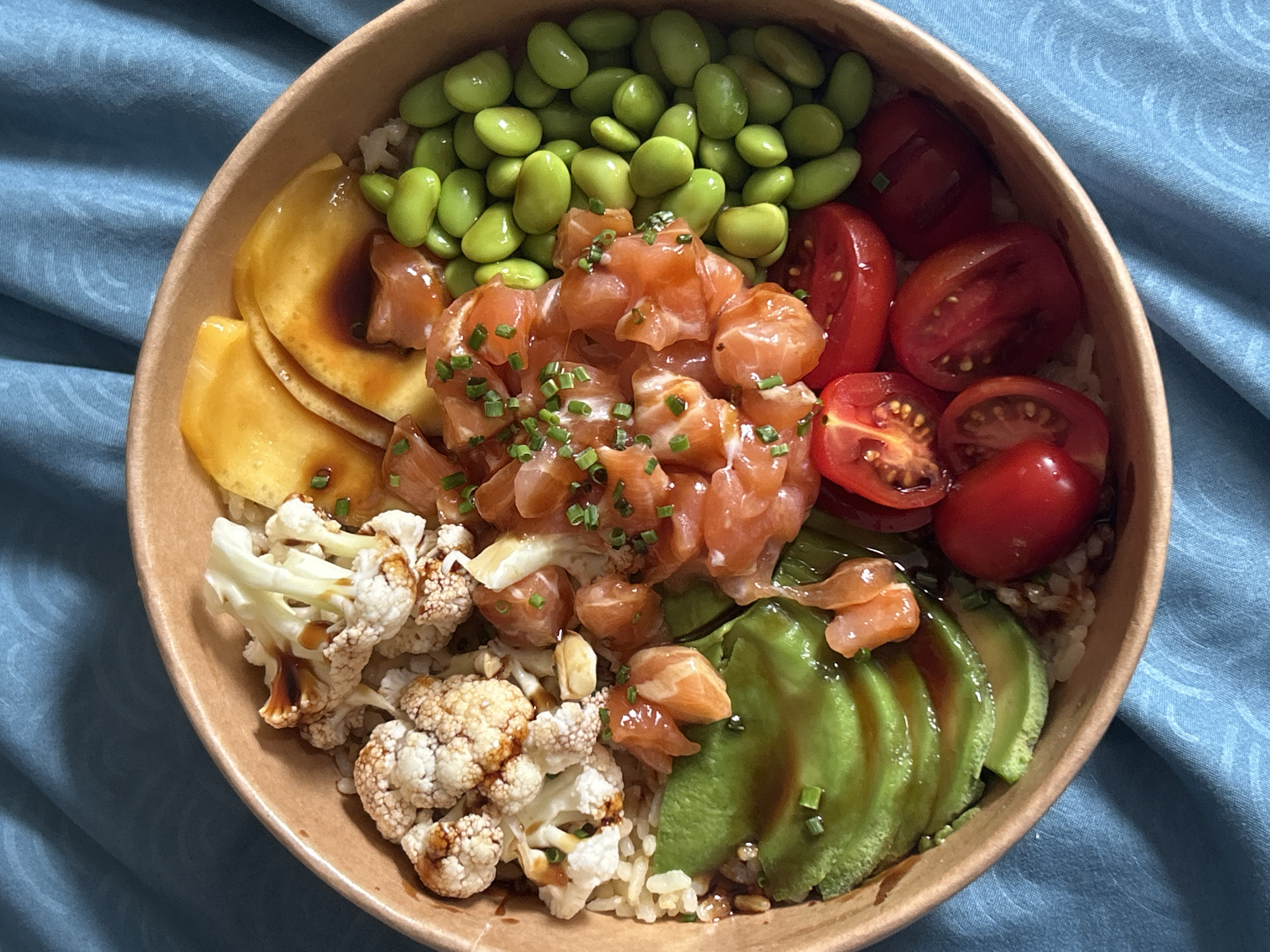 A close-up, overhead shot shows a poke bowl filled with various ingredients. The bowl is round and made of a light brown cardboard material, sitting on a textured blue cloth. Inside, the base appears to be rice, topped with an assortment of colorful elements.
In the center, there's a generous portion of diced raw salmon, glistening and sprinkled with finely chopped green chives. To the left of the salmon, there are several slices of what appear to be mango, with a dark sauce drizzled over them. Above the salmon and mango are bright green edamame beans, arranged in a pile. To the right of the salmon, halved cherry tomatoes are visible, their red skins and juicy interiors showing.
Further to the left and at the bottom of the bowl are pieces of steamed cauliflower florets, some with a light brown char and drizzled with a dark sauce. Adjacent to the salmon and tomatoes, towards the bottom right, are fanned slices of ripe avocado, their green flesh contrasting with the other ingredients.
The lighting suggests it's daytime, and the overall presentation indicates a meal, likely lunch or dinner, being served in a casual setting. The image is sharp and focuses on the food, with no people or specific location cues beyond the blue cloth, which might be a tablecloth or placemat. There is no visible text in the image.