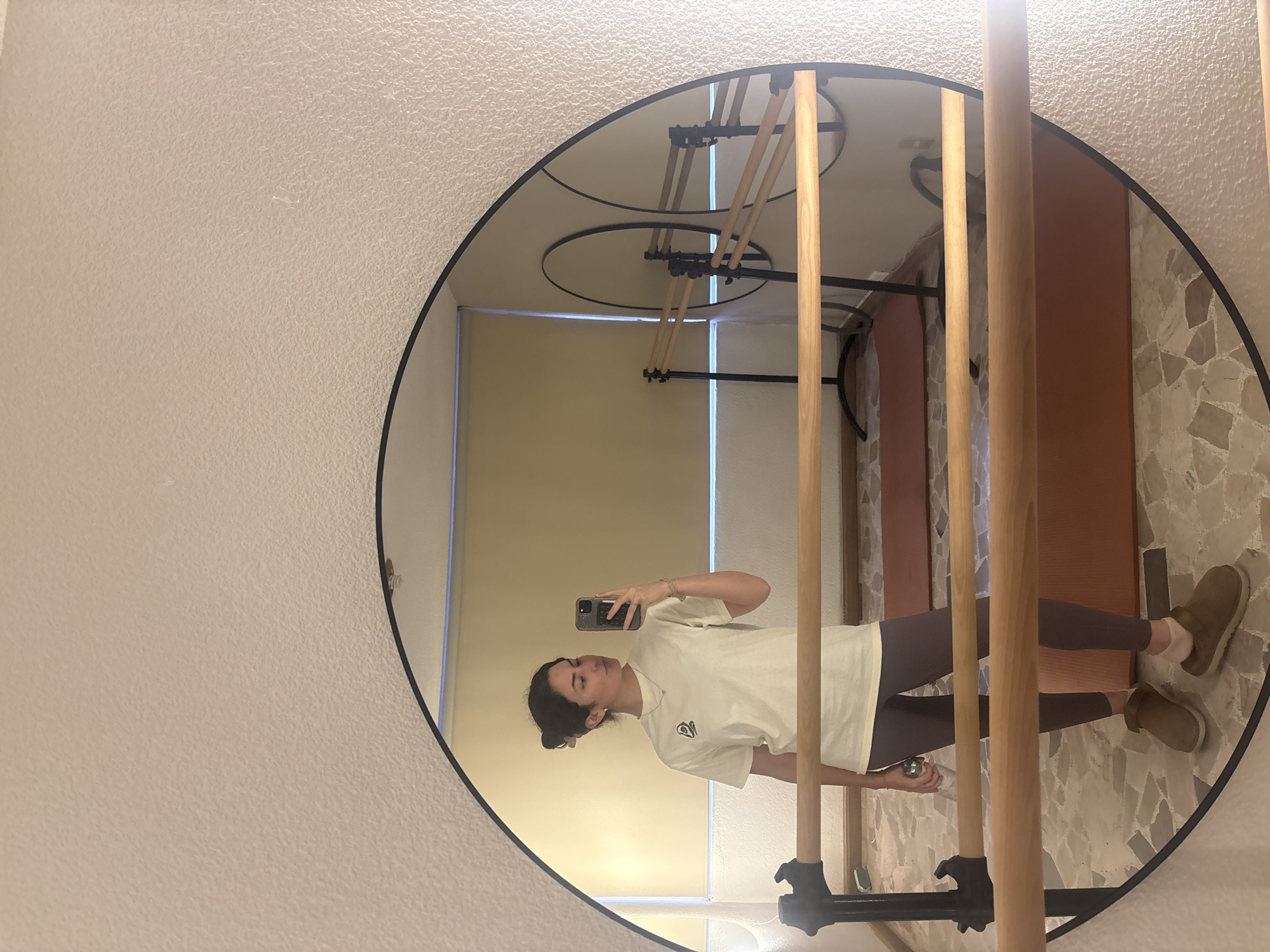 A person stands in front of a round mirror in what appears to be an interior space. The person is holding a smartphone, presumably taking a selfie, and their image is reflected in the mirror. The person wears a light-colored T-shirt, dark leggings, and tan-colored shoes. The environment includes a drying rack visible in the mirror's reflection and a beige tiled floor, suggesting a domestic setting. The photograph is taken at an angle. The location is not evident in the image to determine being in Puebla, Mexico.