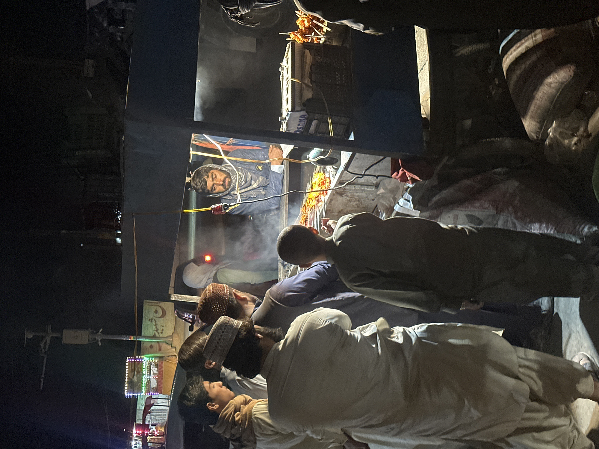 The image shows a group of people gathered around a food stall at night in Quetta City Tehsil, Pakistan. A man is cooking food on skewers, with smoke rising from the grill. Several customers are observing the cooking process, dressed in traditional Pakistani clothing, including hats. The stall is decorated with lights.