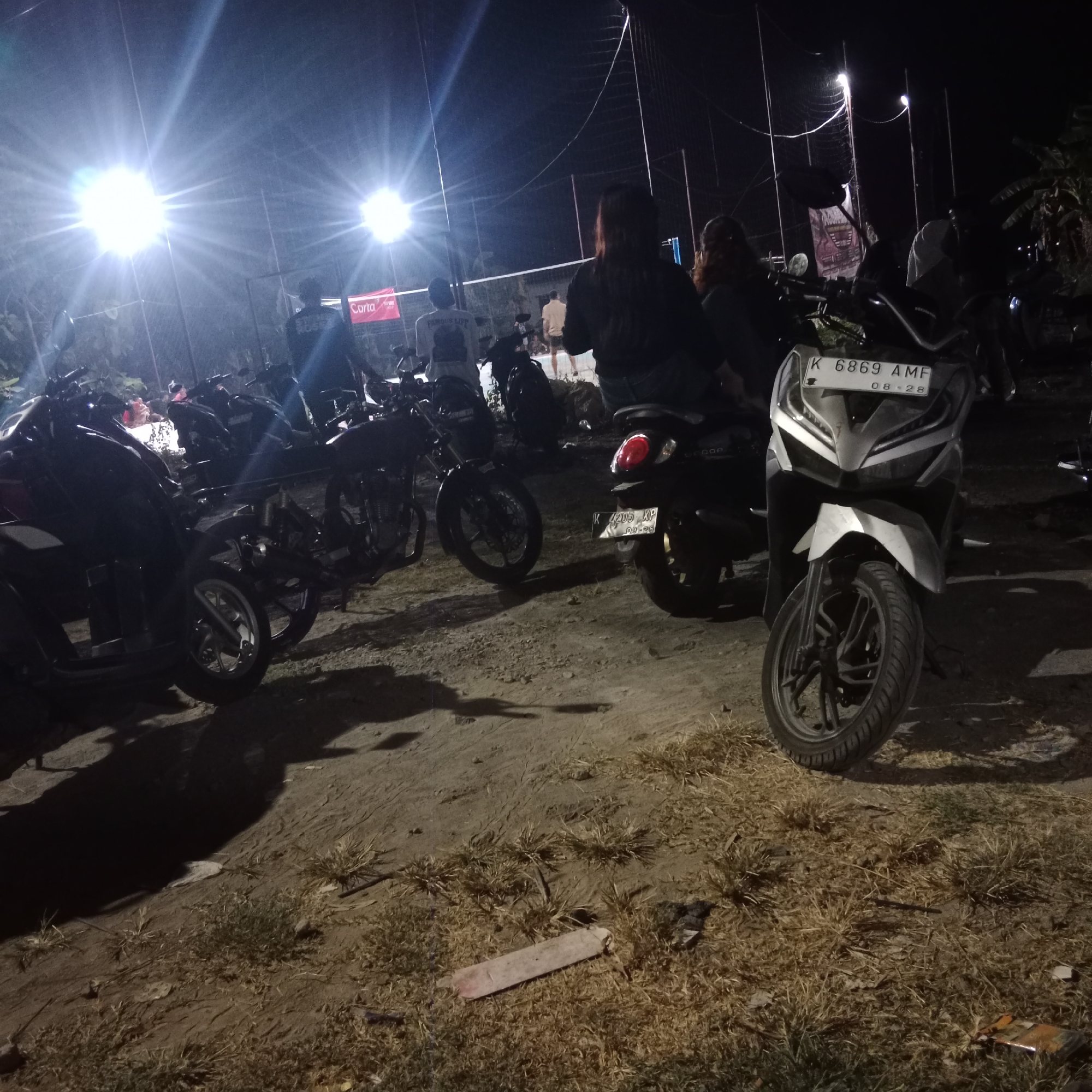 In Kunjeng, Indonesia, a night scene unfolds at a volleyball court illuminated by bright overhead lights, casting strong beams and lens flare. A group of individuals are gathered, with some standing and others on motorcycles. Several motorcycles are parked in the foreground and midground, some with visible license plates, such as "K 6869 AMF" and "K 7405 XP." The ground is a mix of dirt and dry grass. In the background, a volleyball net is visible, suggesting an ongoing or recently concluded game. A red banner with "Carta" written on it is displayed near the court. The overall impression is a casual, outdoor gathering at night.