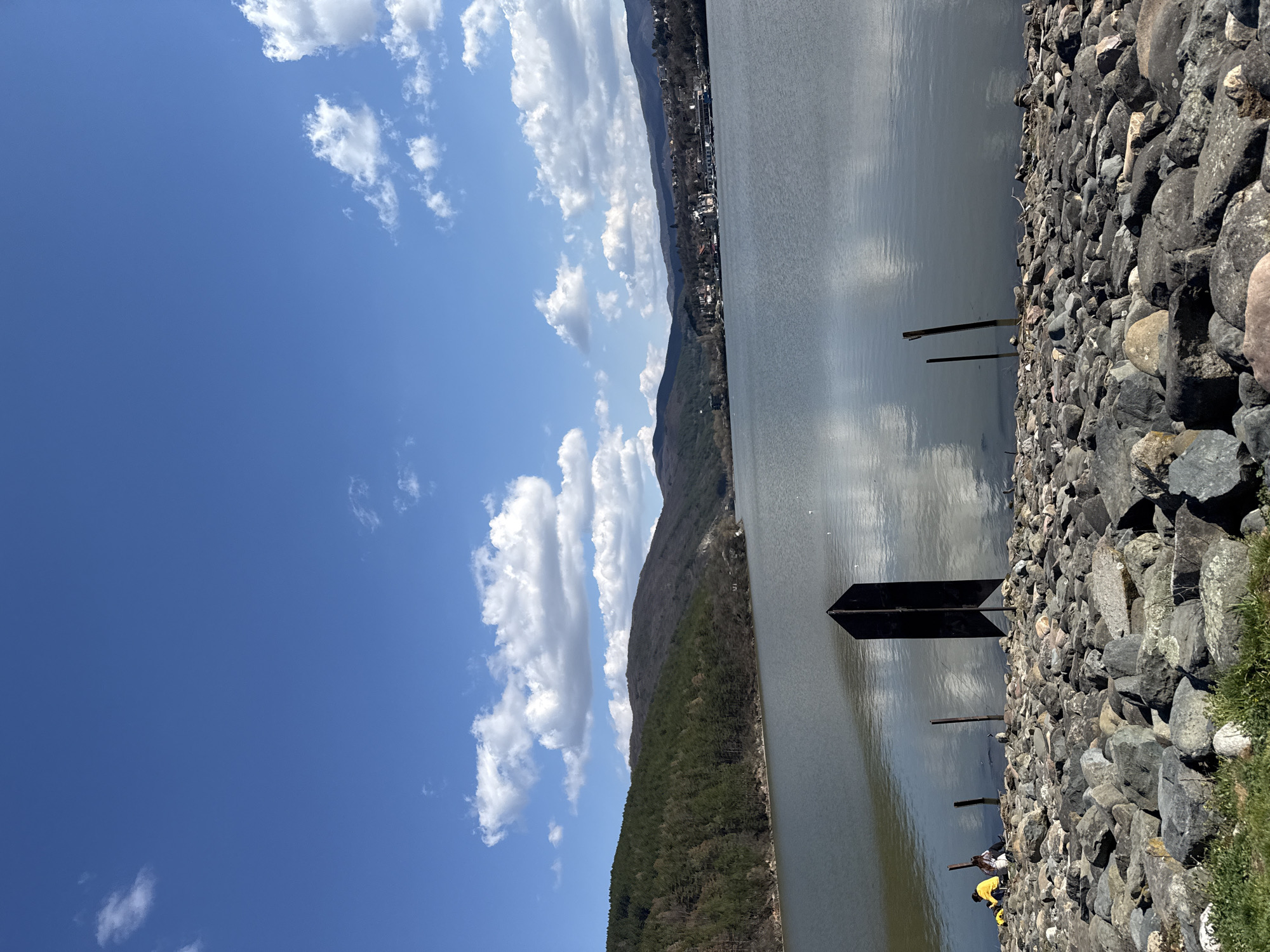 The image depicts a landscape scene featuring a body of water, likely a lake or river, with rocky embankments in the foreground. A metal sign or marker is positioned near the water's edge. The water surface reflects the sky and clouds. In the distance, a mountain range is visible, with a settlement or town stretching along its base. The sky is blue with scattered white clouds. The embankment consists of large rocks of various sizes. The image orientation is rotated, with the sky appearing on the left and the water on the right. The location is unconfirmed as Pancharevo, Bulgaria. It's difficult to ascertain people's actions or interactions based on the image.