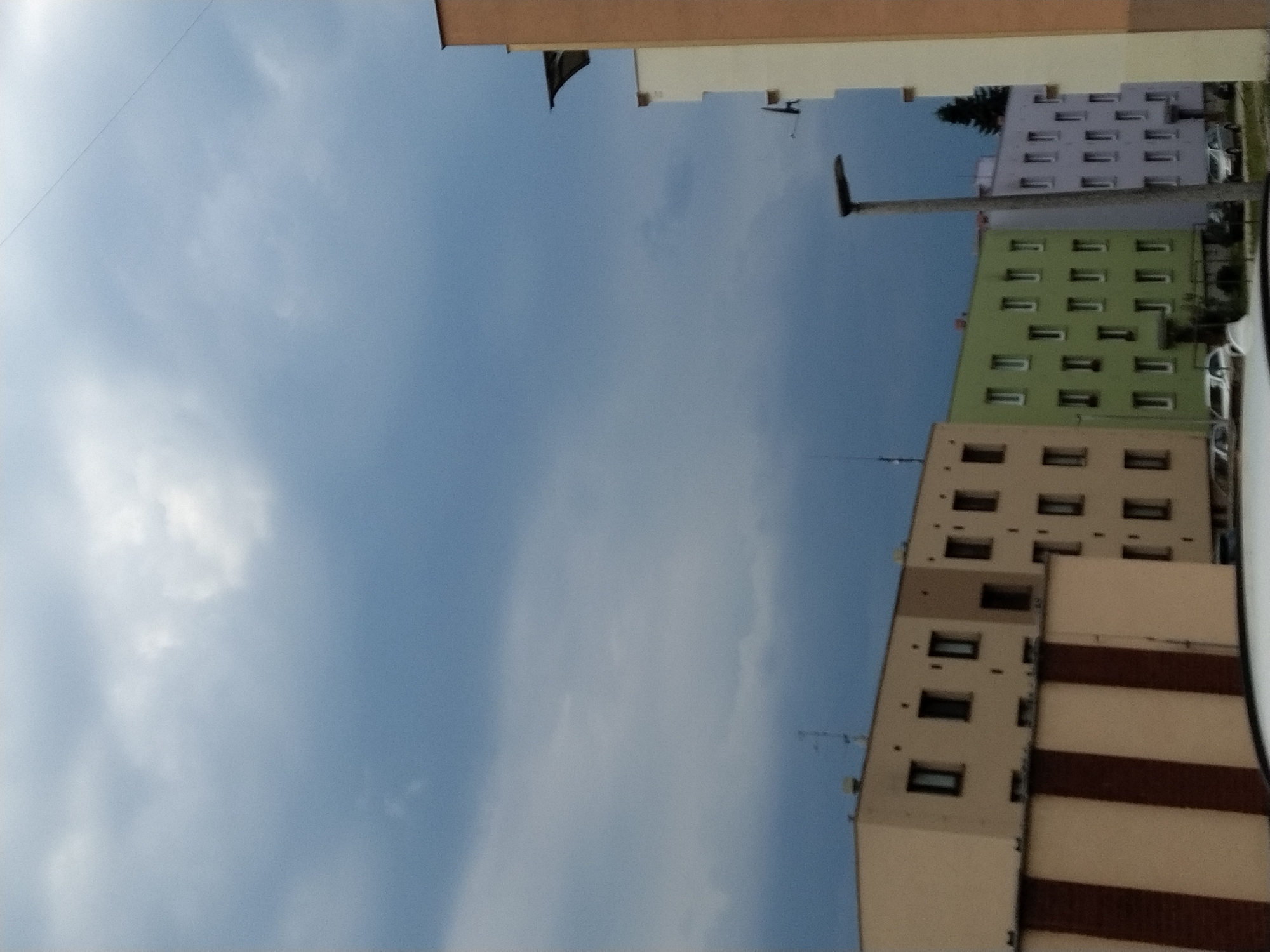 The image depicts a residential street scene in Hluboká nad Vltavou, Czechia, under a cloudy sky. In the foreground, the top of a white car is visible. Adjacent to it, a building with a light brown facade and vertical dark brown stripes is partially visible. Further back, a beige apartment building with multiple windows is lined up. To its right, a light green apartment building with numerous windows is followed by a light purple apartment building, also with many windows. A tall, grey lamppost stands in front of the purple building. In the background, a dark green coniferous tree is visible behind the apartment buildings. The sky is filled with dark, heavy clouds.