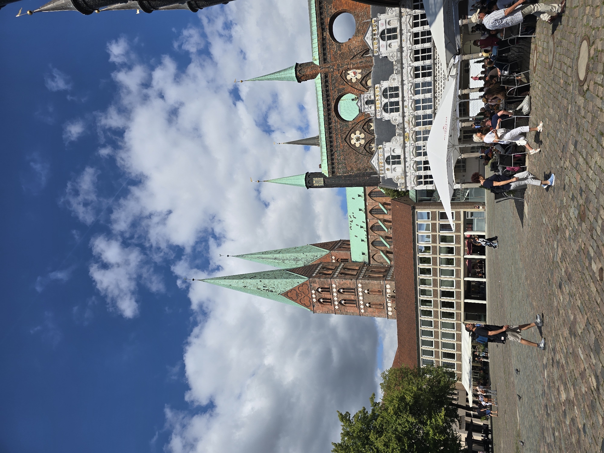 This is a photograph taken from a street-level perspective in the city of Lübeck, Germany, likely in the Markt square. The image features a prominent view of the historic Lübeck Cathedral with its characteristic twin green spires dominating the background. In the foreground and midground, a cobbled plaza is visible, with several people walking and seated at outdoor cafe tables. White umbrellas provide shade over the cafe seating. The architecture in the background includes the cathedral and other brick buildings with distinctive architectural features such as arched windows and decorative elements. The sky is blue with scattered white clouds. The scene depicts a sunny day with moderate activity in a public square.