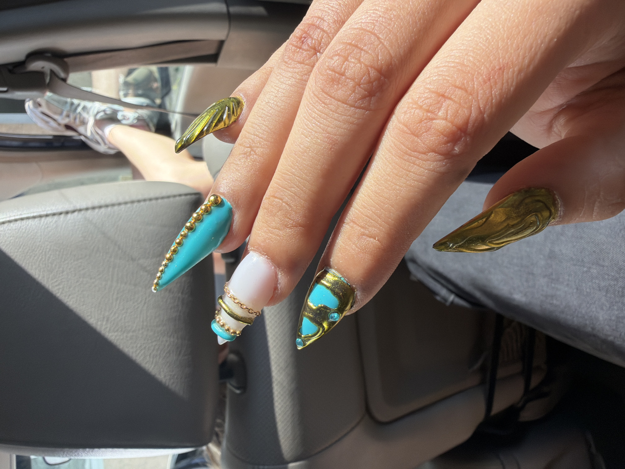 A brightly lit, close-up photograph captures a person's right hand showcasing an elaborate and decorative set of nails within the interior of a car. The hand has a medium skin tone, and the fingers are slightly spread, highlighting each unique nail design.
The nails are long, pointed, and intricately adorned. The pinky and ring fingers feature metallic gold nails with a textured, almost sculpted appearance. The middle finger's nail is white, wrapped with a delicate gold chain and finished with a small turquoise bead at the tip. The index finger displays a solid turquoise nail, detailed with a precise line of tiny gold beads along its outer edge. The thumb nail is metallic gold with two turquoise heart-shaped cutouts and small blue rhinestones.
In the blurred background, elements of a car interior are visible, including grey upholstery and sections of the car's door or dashboard. A person's bare leg, presumably belonging to the same individual, is seen resting in a relaxed position, wearing white socks and grey and white athletic sneakers. Dark grey pants are also partially visible on the right side of the frame.
The image is illuminated by strong natural sunlight, casting clear shadows across the car's interior, suggesting it is daytime. The overall setting implies a casual moment, possibly during travel or while parked. No visible text is present in the image. This scene is set in Burgas, Bulgaria.
