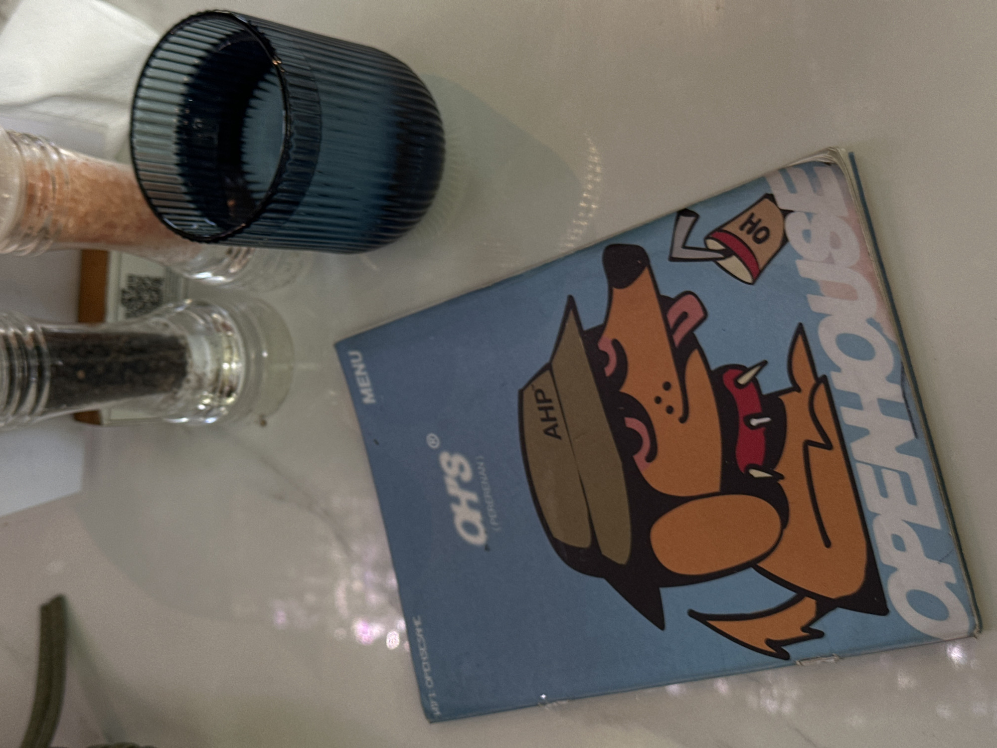 The image is a close-up, eye-level shot of a menu lying on a white, textured surface, likely a table. The menu is light blue and features a cartoon illustration of a dog wearing a hat and sunglasses, holding a drink. The word "OPENHOUSE" is prominently displayed on the bottom of the menu in large, white lettering. The brand "OH'S" is also visible in white lettering above the dog illustration. To the right of the menu is a dark blue, ribbed glass filled with a clear liquid, likely water. Behind the menu and glass, two salt and pepper shakers are visible; one appears to contain black peppercorns and the other pink salt. The background is out of focus, but hints of white fabric and a barcode scanner can be seen. The lighting suggests an indoor setting, possibly a restaurant. The overall scene is clean and organized, with the menu and drink being the central focus. This appears to be a menu from "OH'S OPENHOUSE" located in Mengwi, Indonesia, as indicated by the text on the menu.