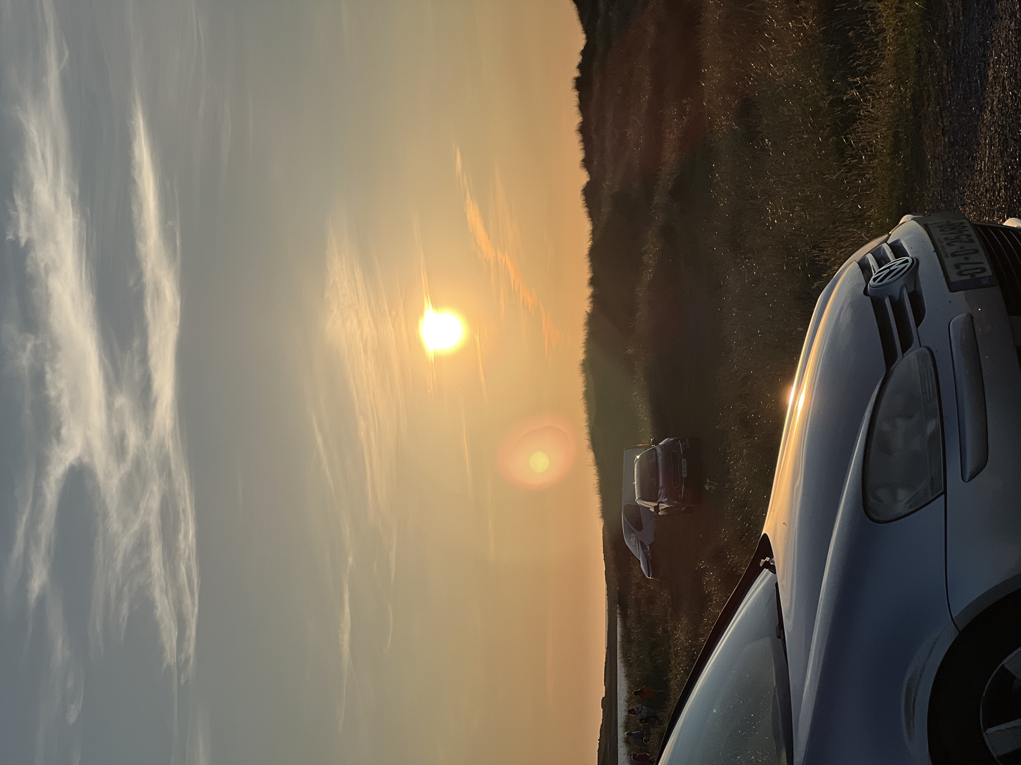 The image captures a sunset over a coastal landscape in Stranacorkra, Ireland. The sun, a bright orange orb, is positioned in the upper center of the frame, casting a warm, golden light across the scene. Wispy clouds are visible in the sky, tinged with the colors of the sunset.
In the foreground, the hood and front of a silver Volkswagen is prominently displayed, reflecting the sunset's glow. The car is parked on a gravel surface.
Behind the car, sand dunes covered in dry grass stretch into the middle ground. Several vehicles are parked amongst the dunes, including a dark-colored car with a visible license plate and a white van.
Further in the distance, on the left side of the image, a body of water is visible, bordering a grassy shoreline. A group of people, appearing as small figures, are gathered near the water's edge. They are casually dressed and engaged in unspecified activities.
The overall impression is one of a peaceful evening at the coast, with the natural beauty of the sunset enhanced by the presence of people and vehicles. The image seems to have been taken from a slightly elevated position, looking across the landscape.