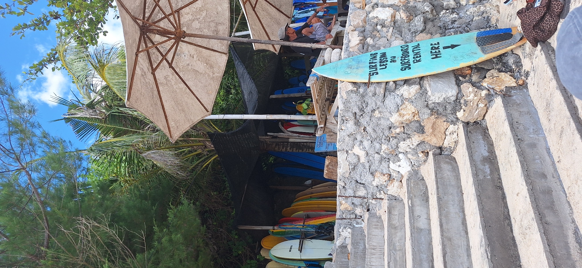 The image depicts a surf school and rental stand located in Kuta Selatan, Indonesia. In the foreground, a turquoise surfboard with black lettering stands upright against a stone wall. The lettering on the surfboard reads "SURFING LESSON AND SURFBOARD RENTAL HERE" with an arrow pointing downwards. A set of concrete stairs leads up to the left of the surfboard.
Behind the stone wall and surfboard, a collection of surfboards of various colors and sizes are propped up and displayed under a shaded canopy. Palm trees and other lush vegetation are visible in the background, suggesting a tropical environment. Two large, light-colored beach umbrellas provide shade over seating areas.
A man, wearing a grey cap, dark shirt, and light-colored shorts, is seated on a low bench or table on the right side of the image, looking to the right. He appears to be an employee or a patron of the surf school. Some clothing items are visible on the ground near the man. The overall scene suggests a relaxed, beachside atmosphere focused on surfing activities.
