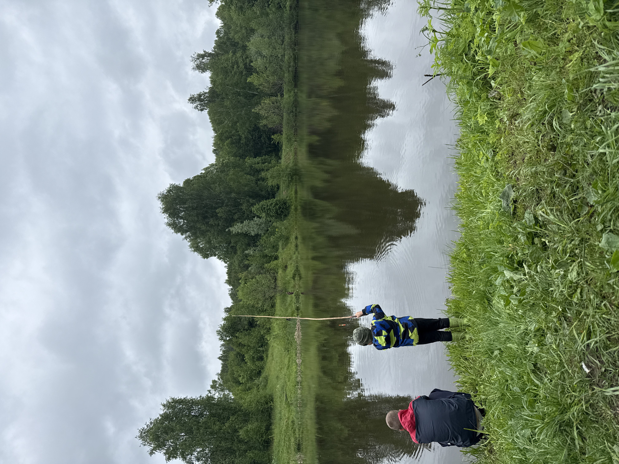A low-angle, outdoor, eye-level shot depicts two people, presumably fishing, on the bank of a body of water, possibly in Zhigalki, Russia. The scene is dominated by an overcast sky reflected in the water. Lush green trees line the opposite bank and are also reflected in the water. The person on the left is holding a long, thin fishing rod, while the person on the right is crouched. The immediate foreground is filled with tall, green grass. The overall tone is tranquil.