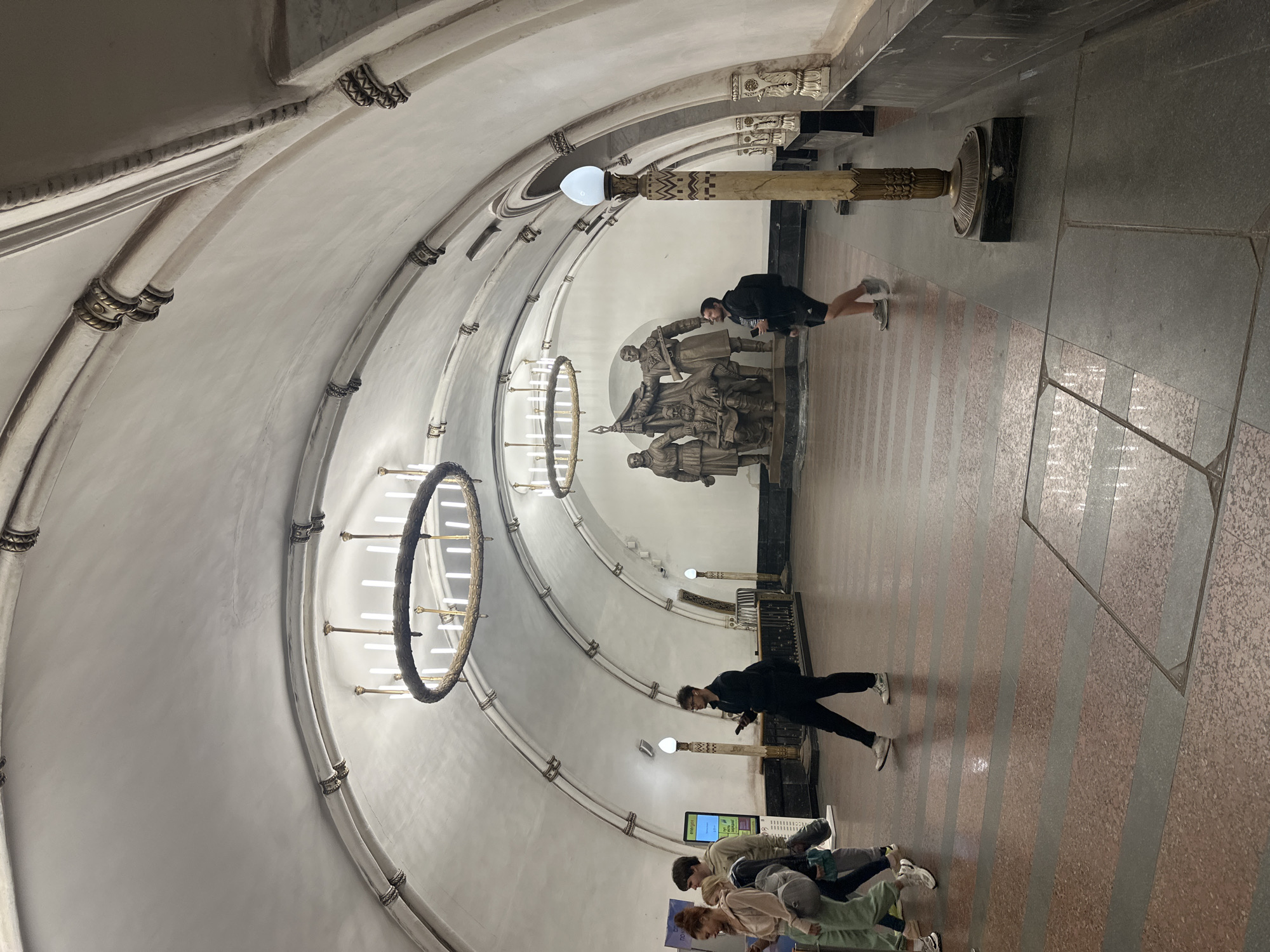 The image depicts the interior of a grand, ornate subway station with a high, arched ceiling adorned with decorative moldings and large, circular chandeliers. Several people are walking through the station, some singly and some in small groups. In the background, a large bronze sculpture of several figures stands prominently. The floor is made of polished stone with alternating bands of red and gray. A tall, decorative lamp post with a glowing orb stands to the right of the frame. The overall atmosphere is one of historical grandeur and public transit.
