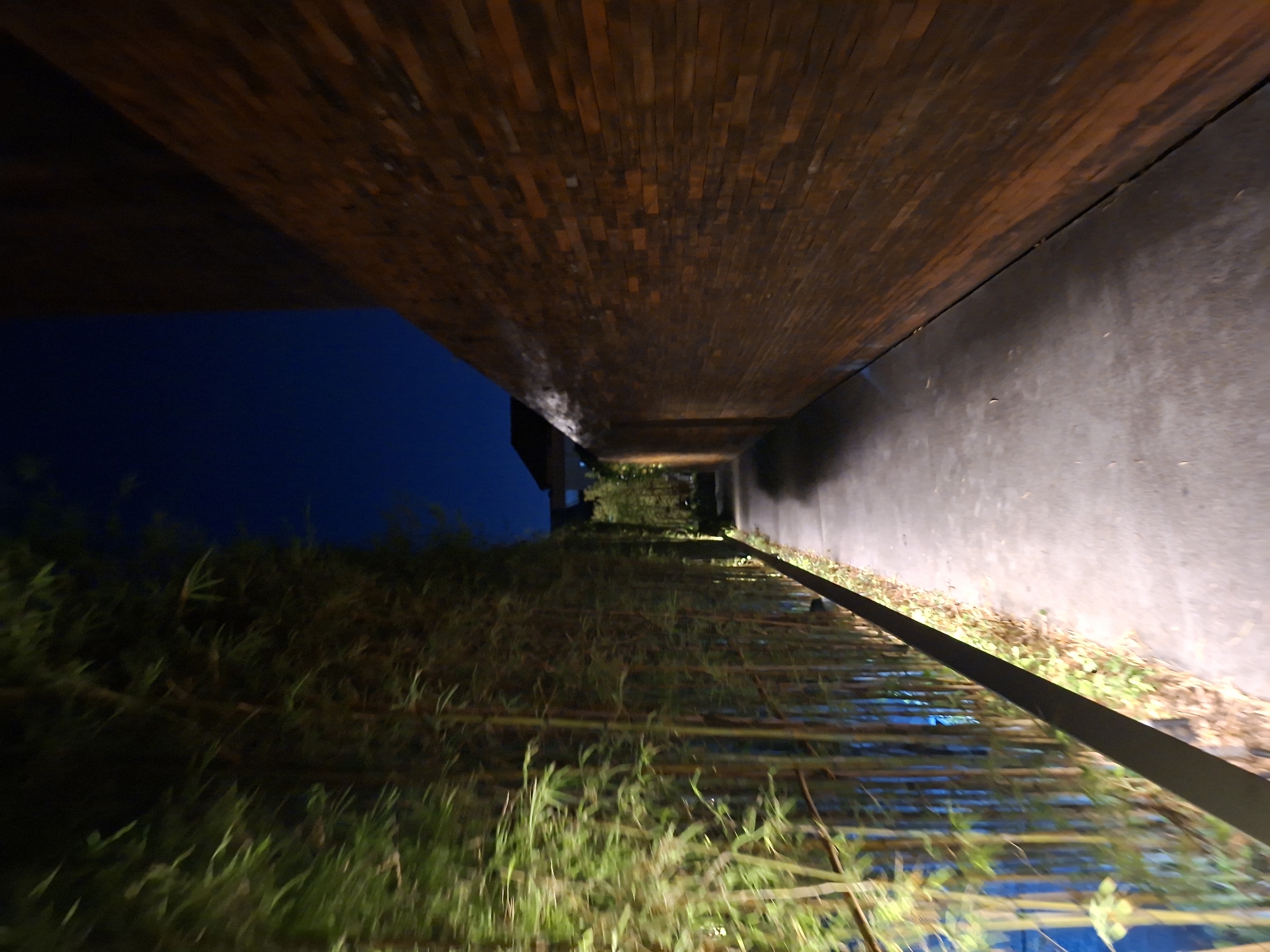 A narrow, paved pathway stretches into the distance, bordered on the left by a dense wall of bamboo illuminated by soft green uplighting, and on the right by a tall, dark brick wall that recedes into shadow. The sky above is a deep, clear blue. The scene appears to be an outdoor passage at night, possibly within a resort or private property. The bamboo on the left is lush and vibrant, its stalks and leaves catching the light, creating a sense of enclosure. The brick wall on the right is made of large, rectangular bricks laid in a horizontal pattern, giving it a textured and imposing appearance. The pathway itself is smooth and grey, with a slight sheen suggesting it might be damp or recently cleaned. A thin, dark line runs along the edge of the pathway, separating it from the bamboo. In the far distance, the roofline of a building is visible against the dark sky. The overall impression is one of a serene and somewhat mysterious passage, designed to be both visually appealing and to guide visitors through the landscape. The location is likely in North Kuta, Indonesia, given the common use of bamboo and the architectural style that can be found in that region.