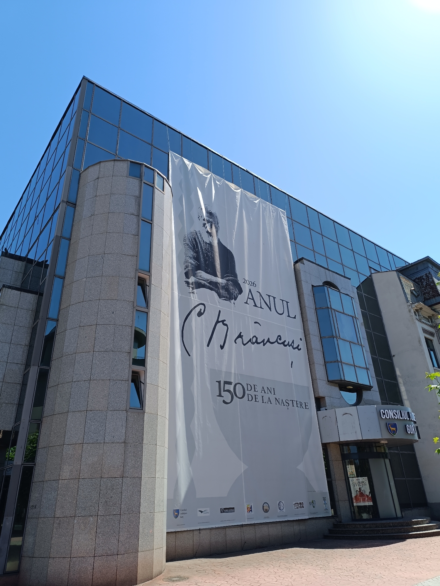 The image depicts a large banner hanging from the facade of a modern building in Târgu Jiu, Romania. The banner features a black and white portrait of a man, presumably Constantin Brâncuși, with text indicating "2026 ANUL BRÂNCUȘI" and "150 DE ANI DE LA NAȘTERE" (150 Years Since Birth). The building itself has a contemporary design with a combination of glass and stone cladding. Below the banner, the text "CONSILIUL JUDETEAN GORJ" is visible, identifying the institution as the Gorj County Council. The scene is set against a clear blue sky on a sunny day.