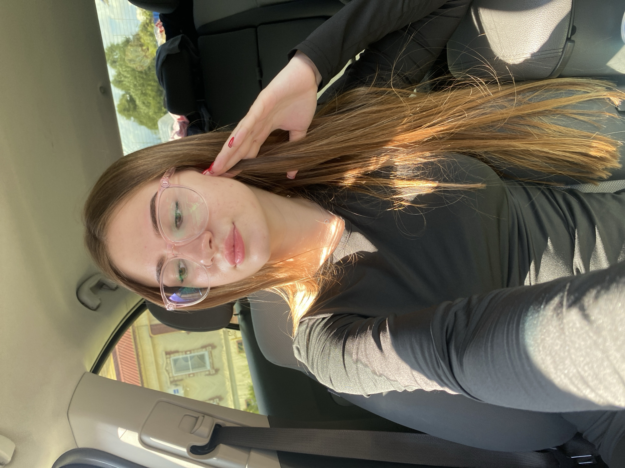 A young woman with long, light brown hair and pink-framed glasses is sitting in the passenger seat of a car, which is parked on a street in the city of Dyakovo, Russia. The woman is wearing a black, long-sleeved shirt. Her right hand is touching her hair, near her ear. Sunlight is illuminating her hair and arm, creating shadows on her clothing. Outside the car, a building with a red roof and a window is visible. The interior of the car is dark, with black seats.