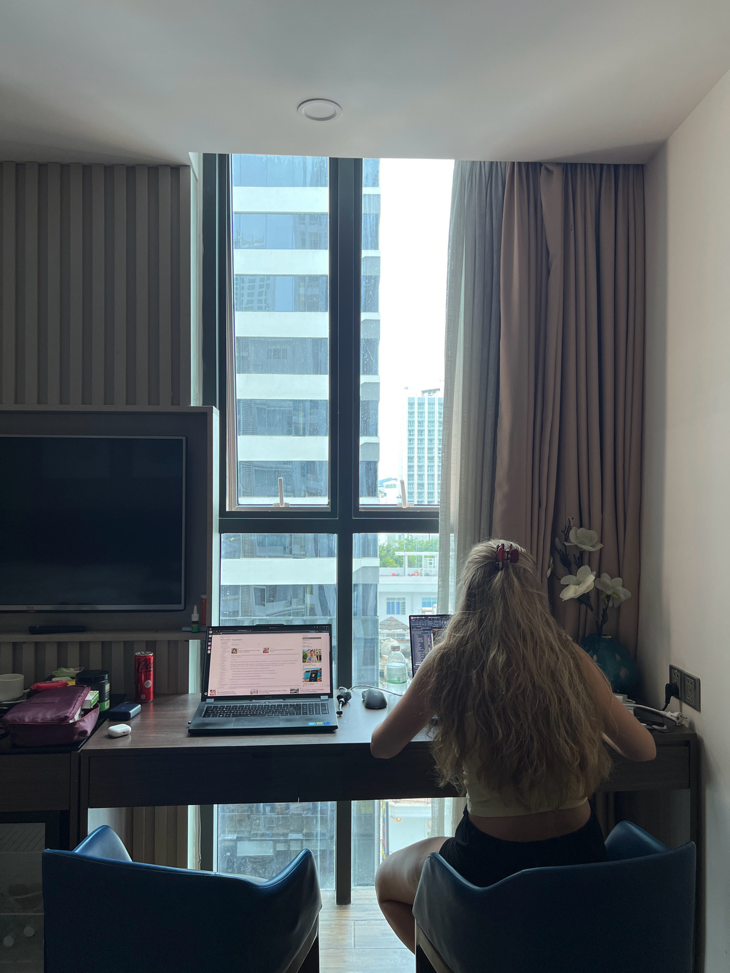 A woman with long blonde hair tied with a red clip sits at a desk in a modern apartment in Nha Trang, Vietnam, facing away from the camera and toward a laptop. She is wearing a yellow tank top and black shorts. The desk is made of dark wood and holds a laptop, a computer mouse, a bottle of water, and a can of Coca-Cola. To the left of the desk is a large television screen and some shelves. Behind the desk, a large window offers a view of tall buildings in the city. The room is well-lit by natural light coming through the window, and the curtains are drawn partially open.